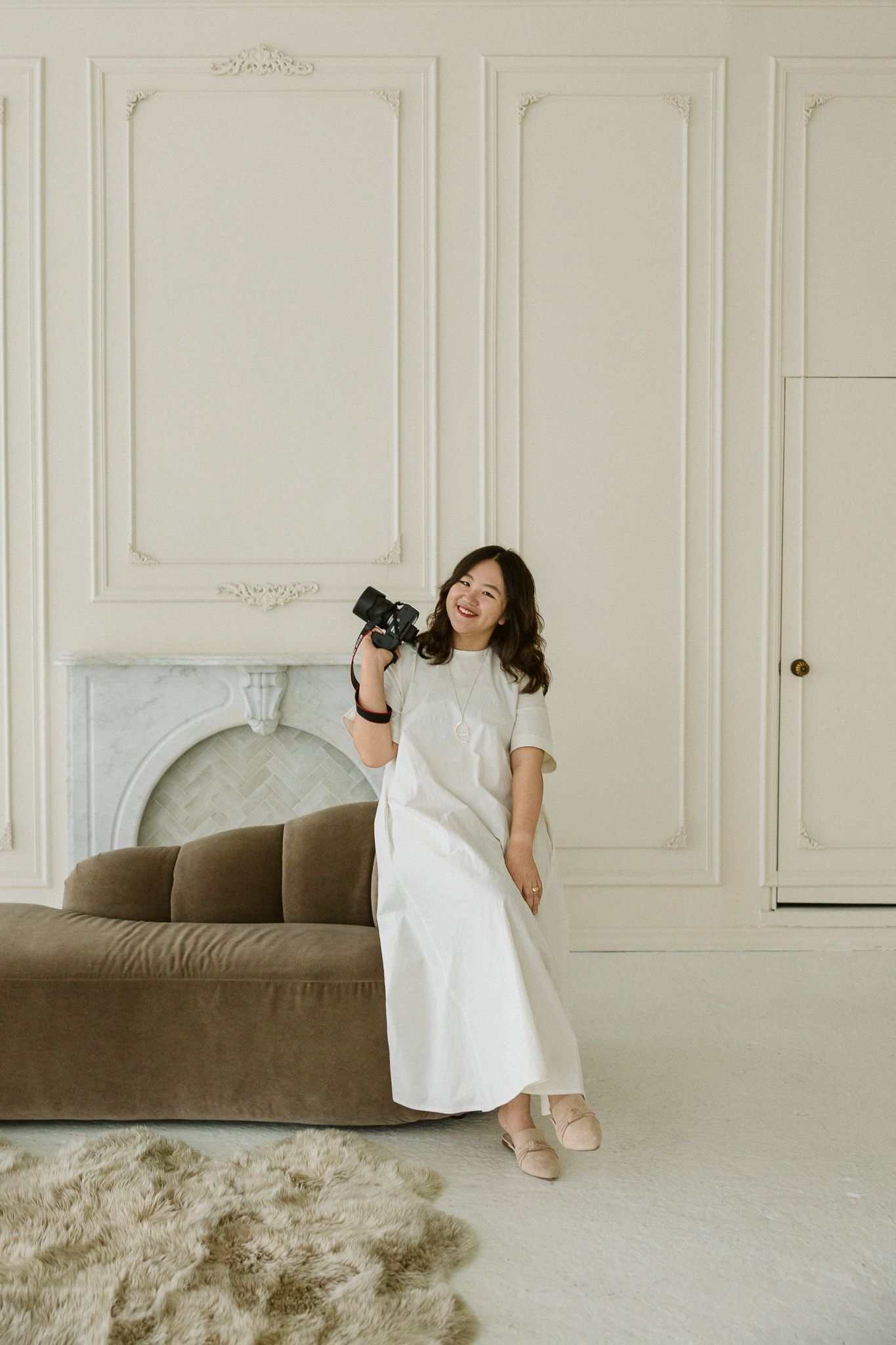 Smiling woman in white dress holding a camera stands in an elegant white room with marble fireplace and velvet sofa.