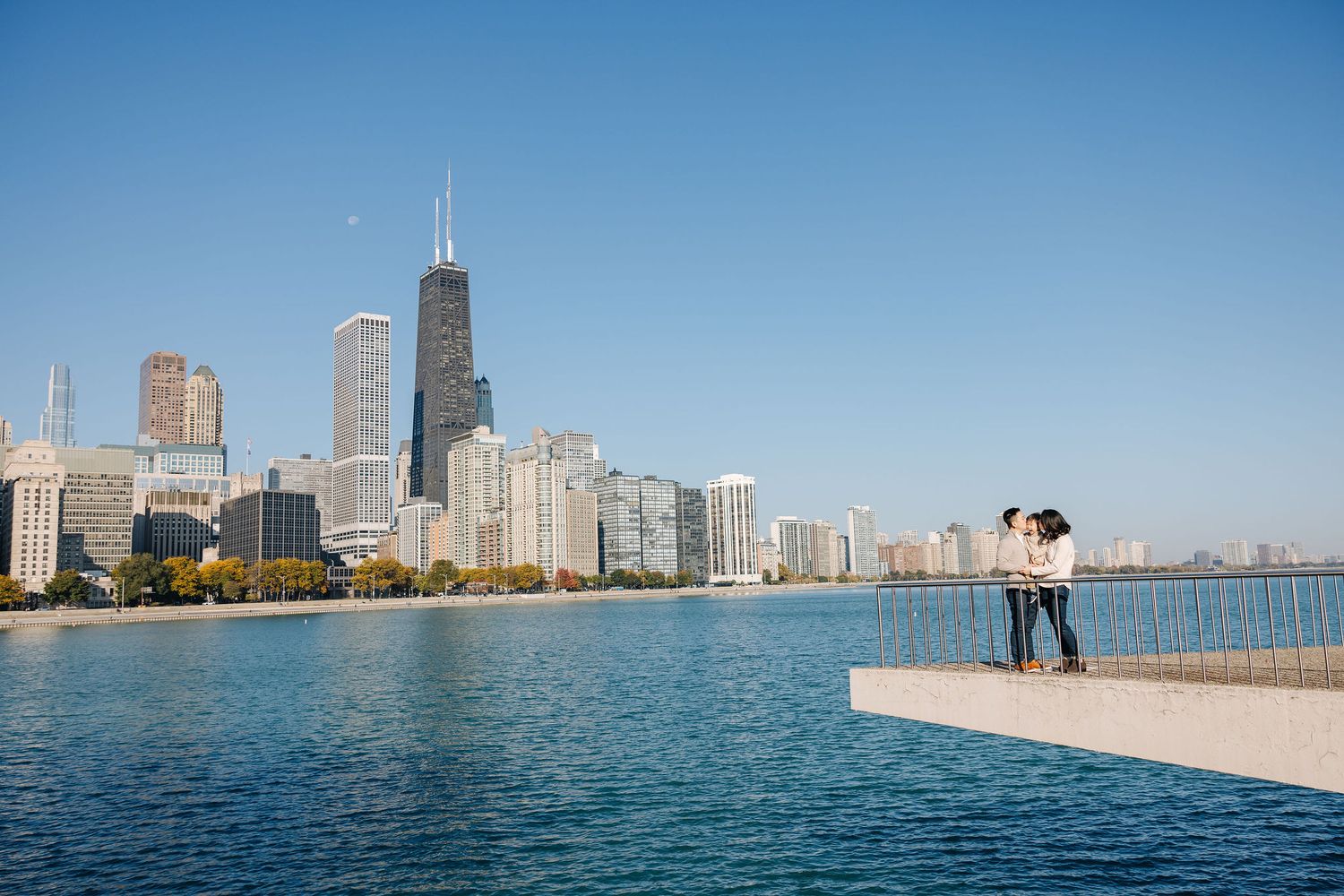Couple embracing on Chicago lakefront with stunning city skyline and John Hancock Center in background on clear blue day.