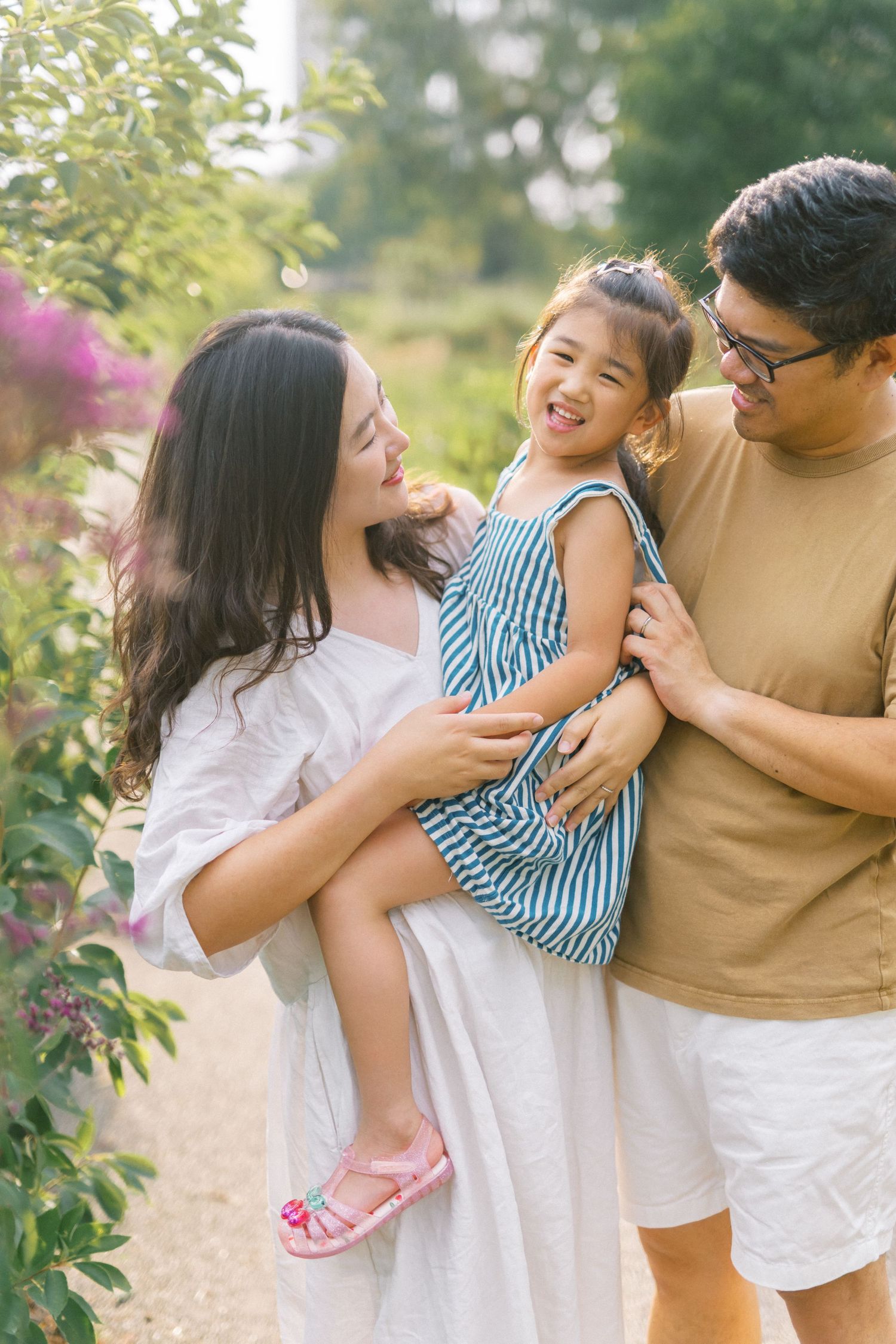 Happy Asian family with parents holding laughing toddler girl in striped dress outdoors in garden.