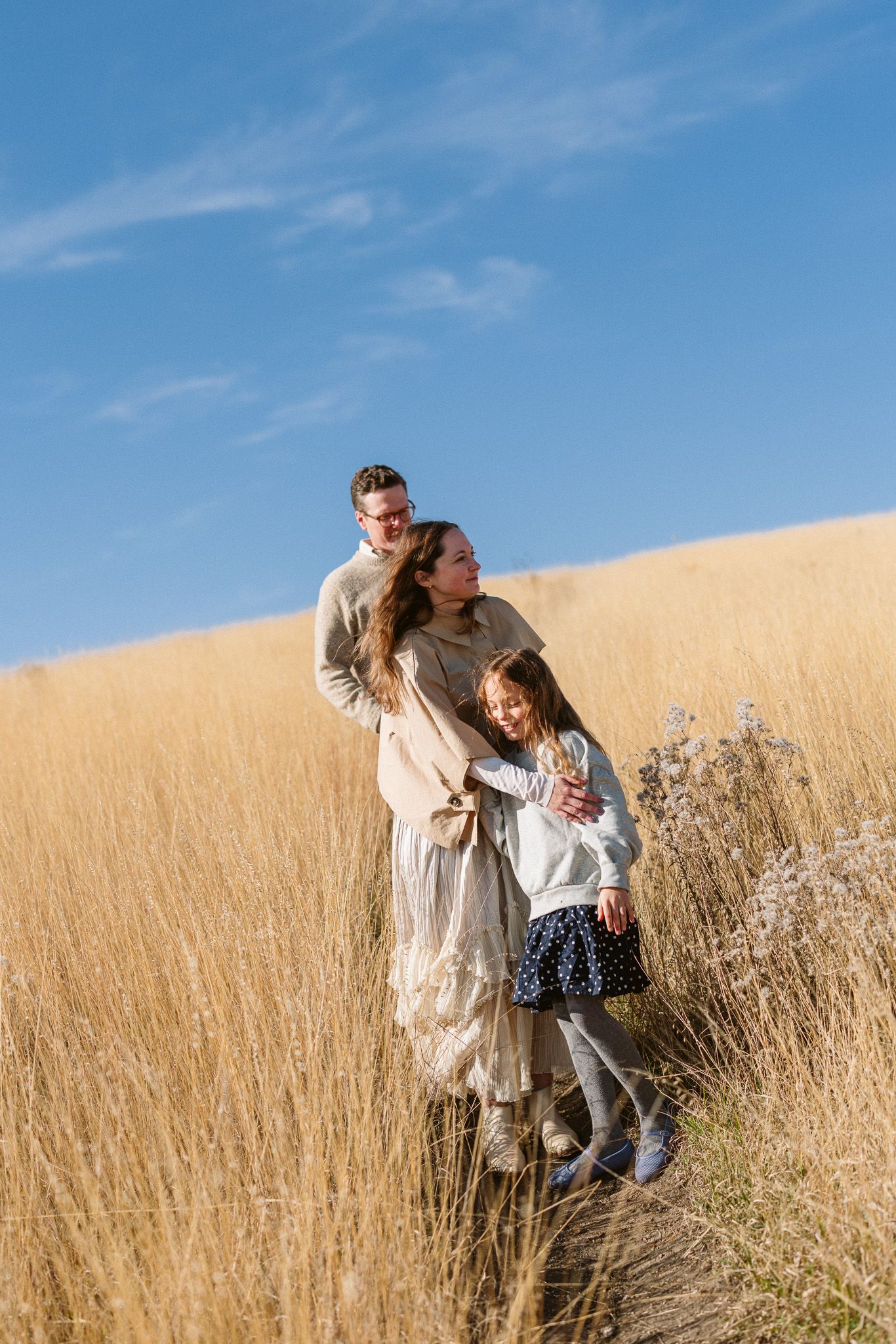 Family of three standing in golden grass field under blue sky, parents embracing child on sunny day.