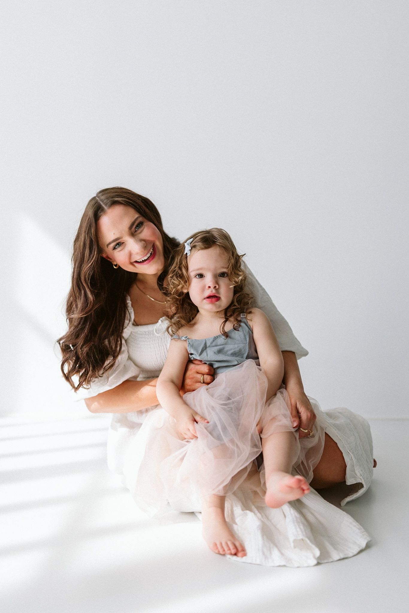 A heartwarming studio portrait of a parent and child in soft white dresses sitting together on a light backdrop.