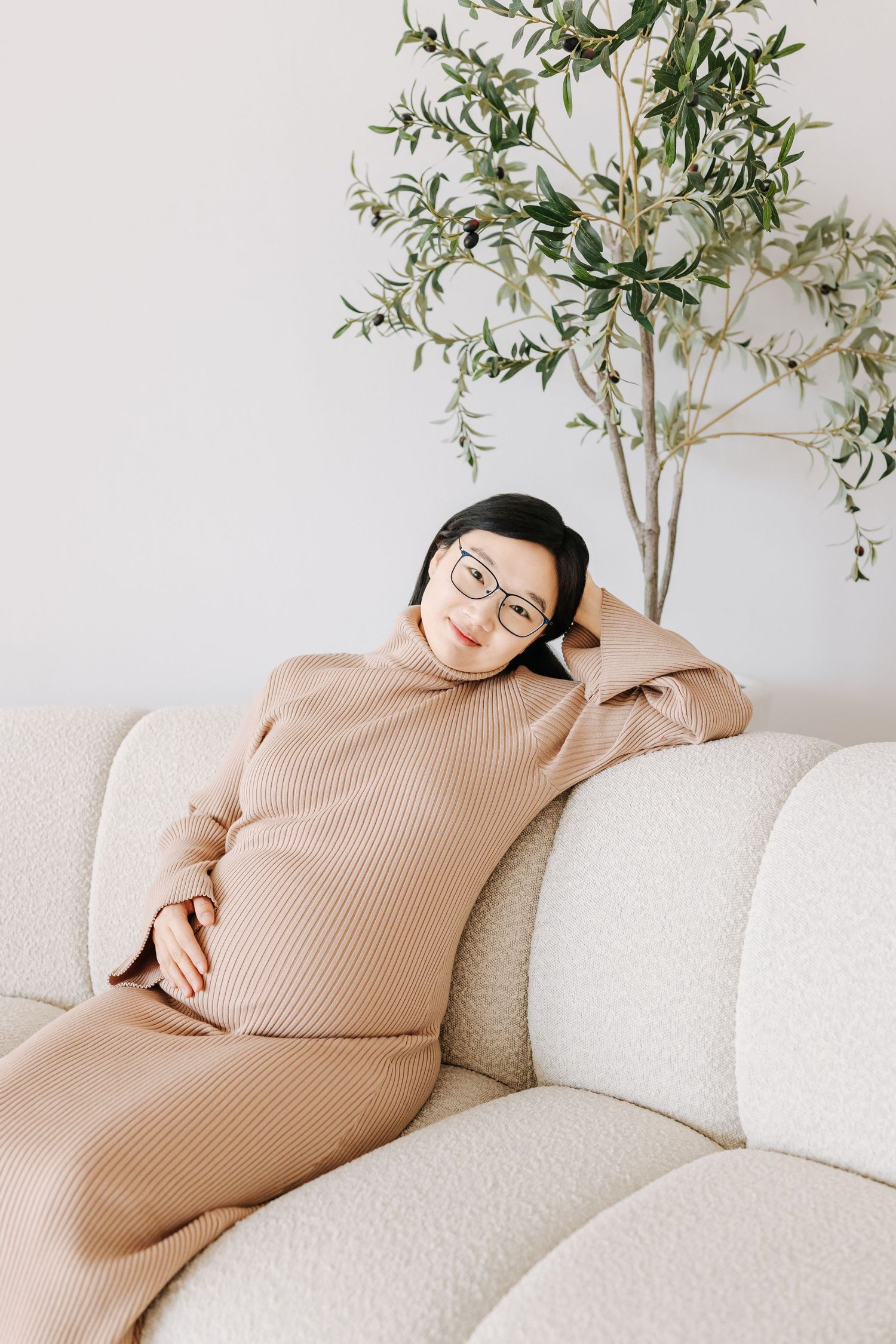 Expectant mother relaxing on white couch wearing beige sweater with indoor plants in background.