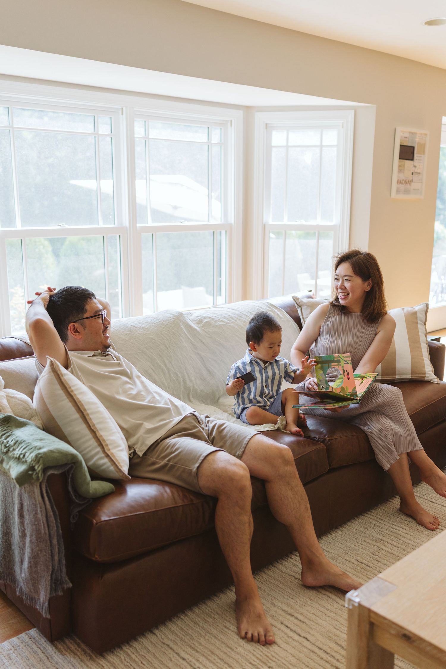 A family relaxes together on a brown leather couch in a sunny living room with large windows.