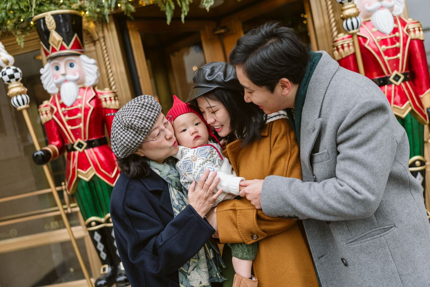 Family in winter coats gathers around a smiling baby in a red hat beside festive nutcracker soldier statues at a holiday display.