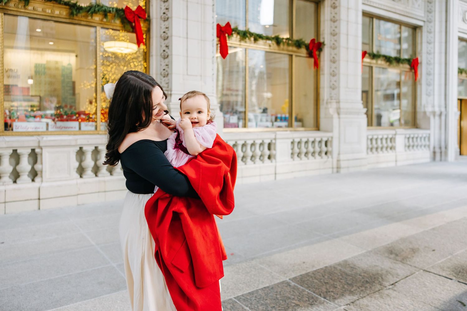 Mother in a red coat holds her baby outside a building decorated with red bows and holiday garland on a sunny city plaza.