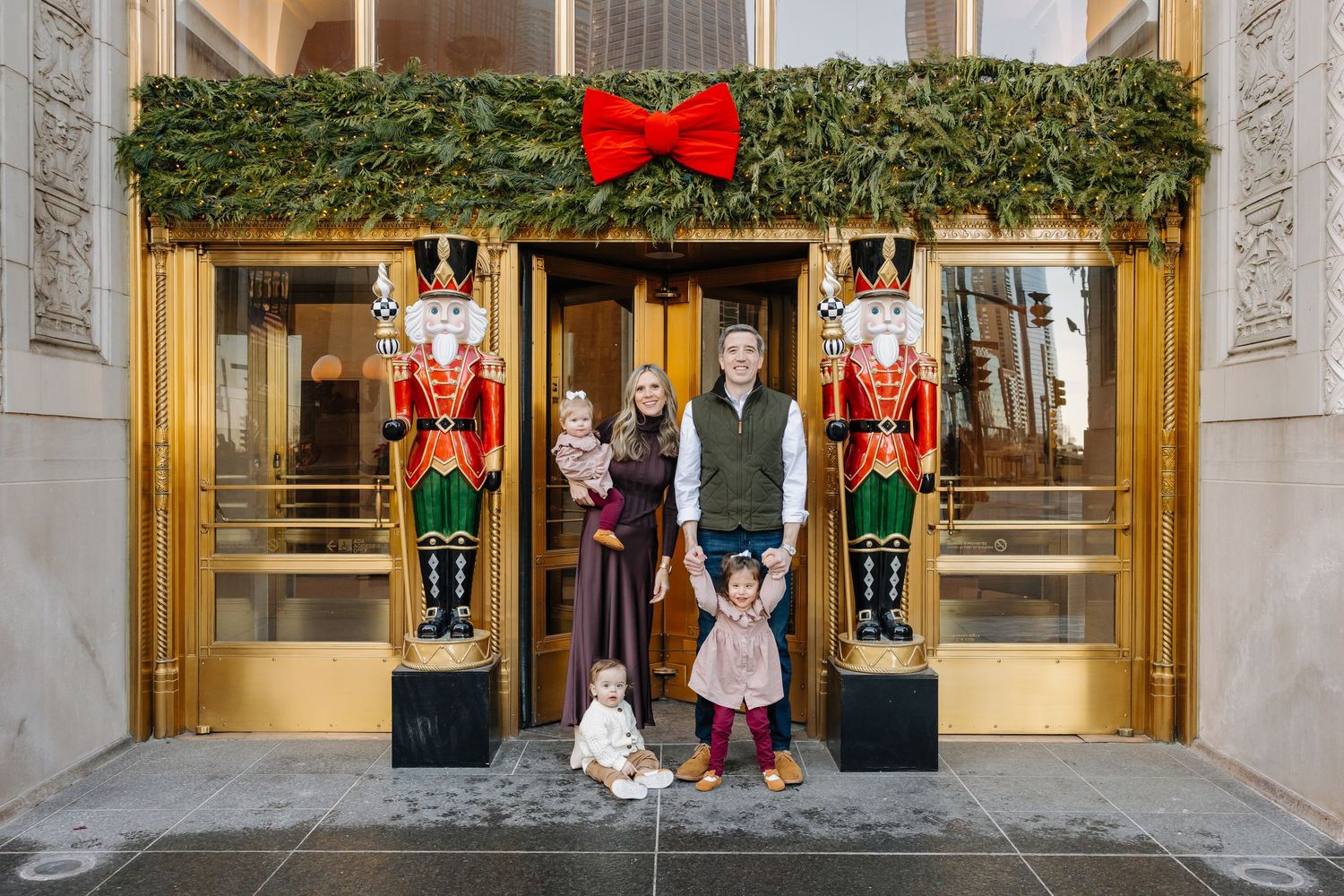Family of five poses in front of golden doors decorated with a red bow, evergreen garland and two life-size nutcracker soldier statues.