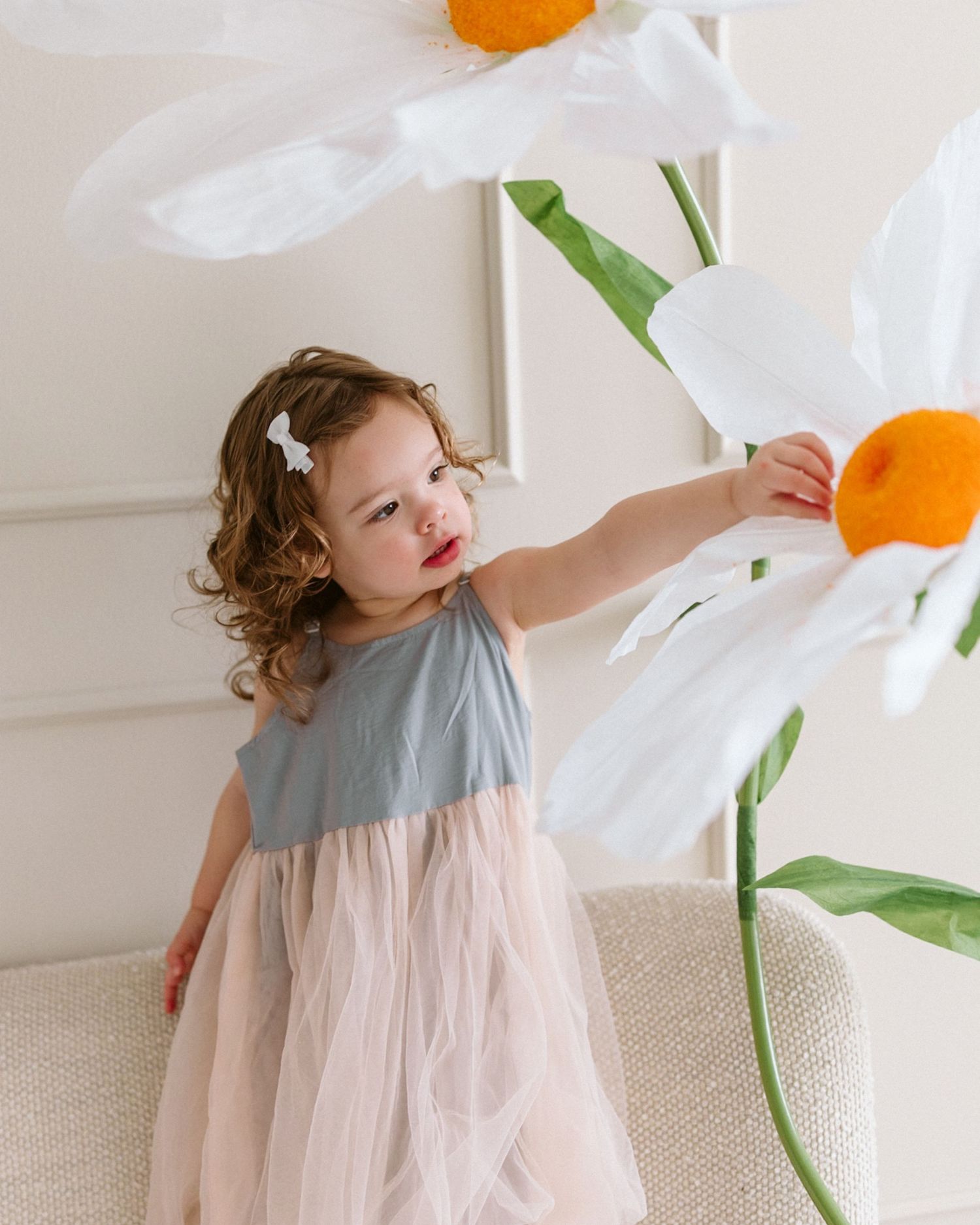A child in a blue and pink tulle dress reaches up to touch large white and orange paper flower decorations.