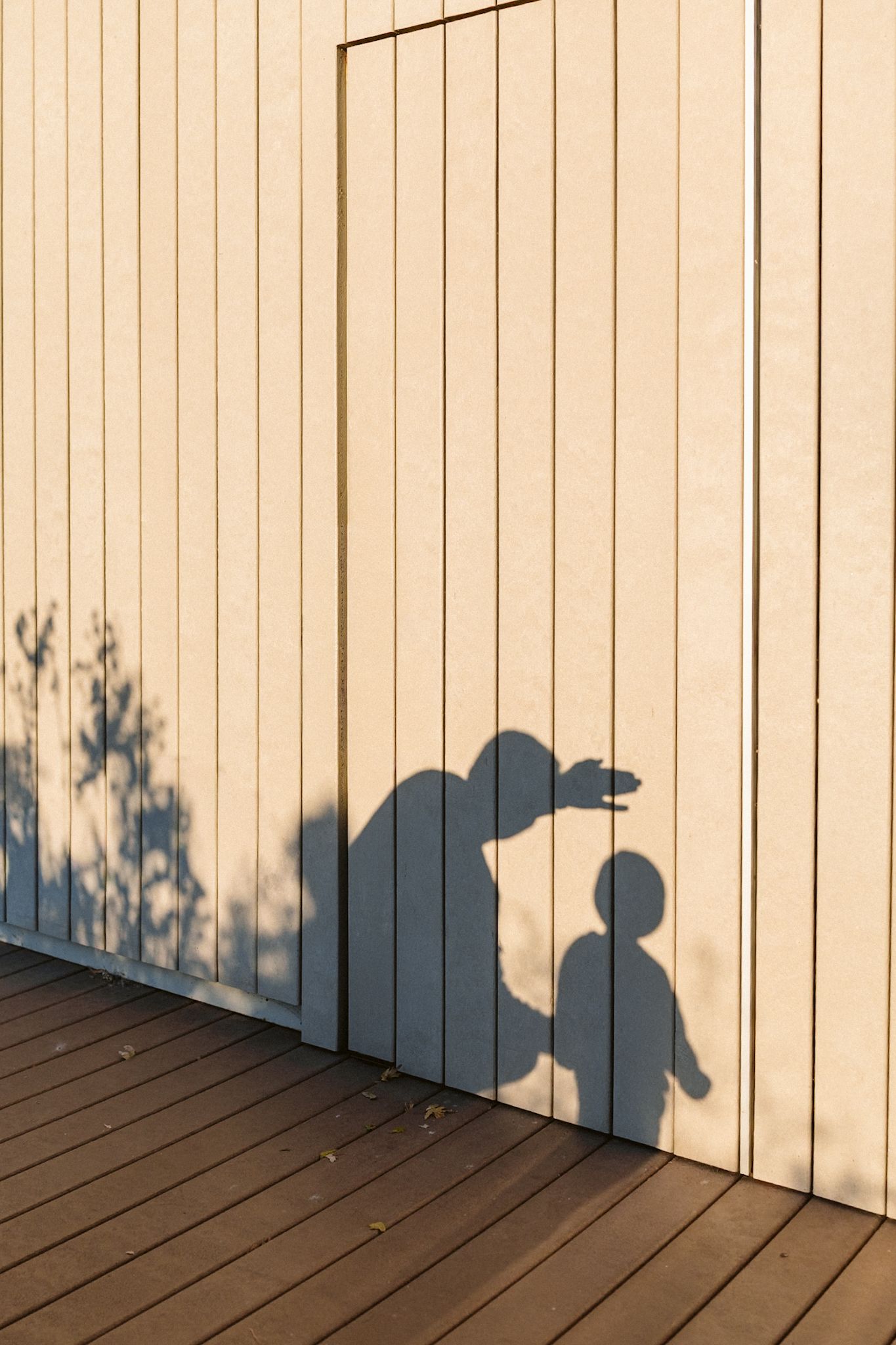 Shadow of adult bending toward child cast on cream vertical wood panel wall above brown wooden deck floor.