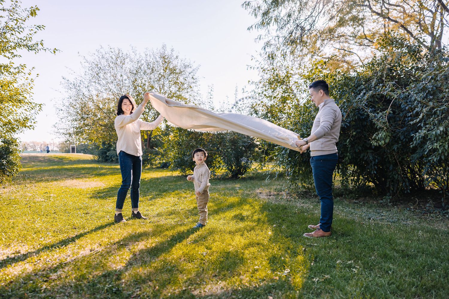Family spreading a blanket on green grass in a sunny park, with a young child standing between two adults.