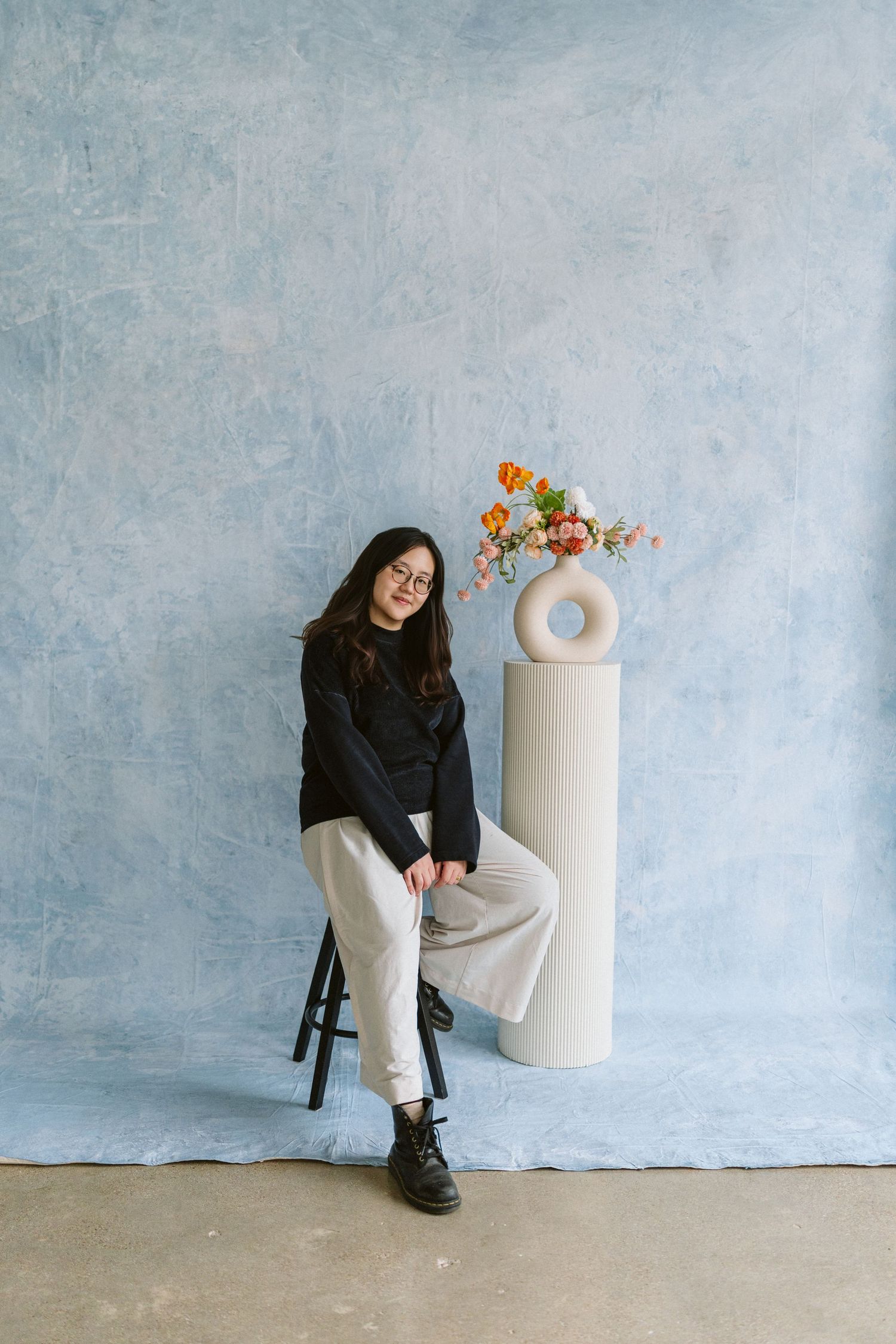 Minimalist studio setup with a cylindrical white pedestal holding orange flowers against a textured blue-gray wall.