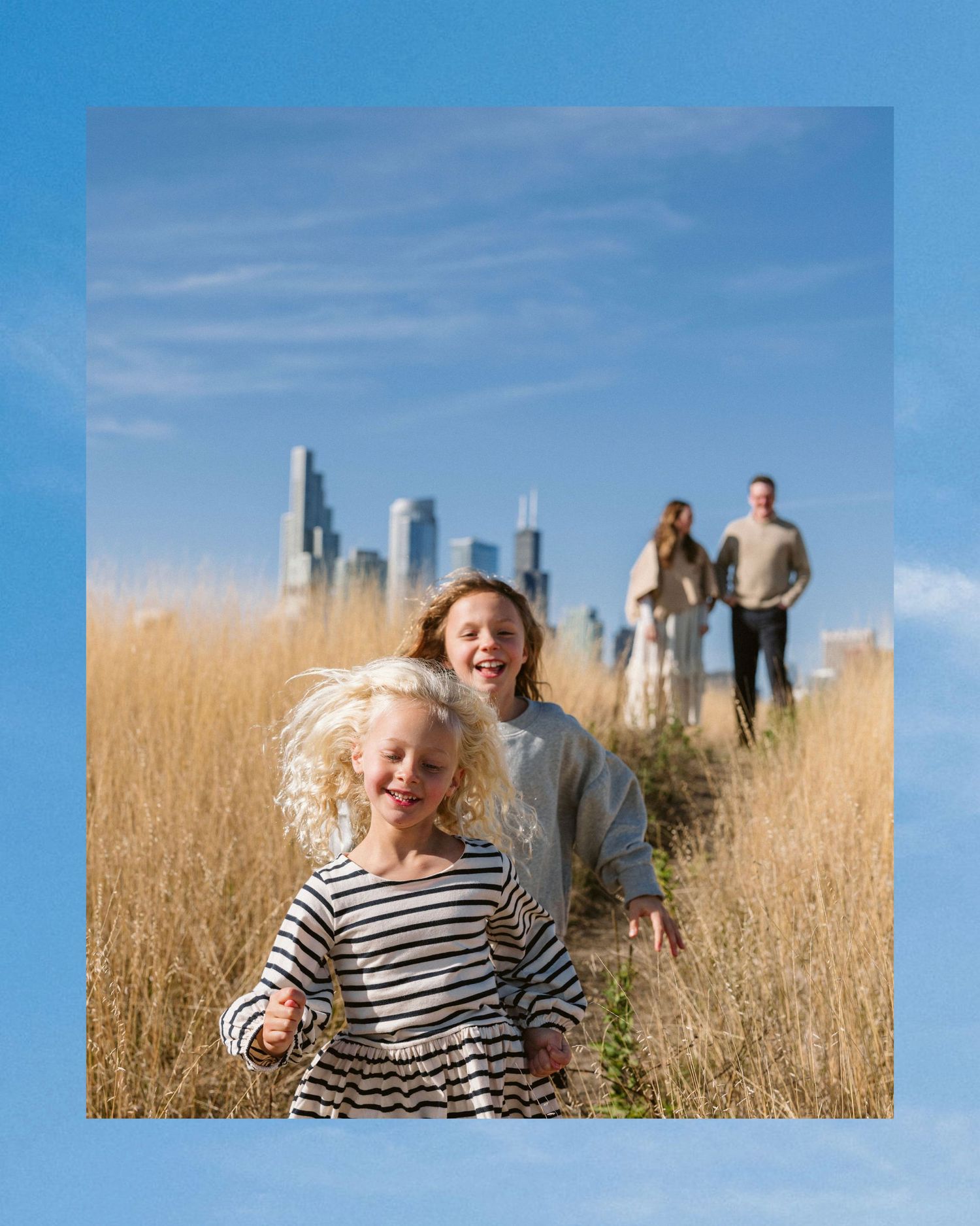 Two blonde girls run through tall golden grass with parents and city skyline in background on sunny day.
