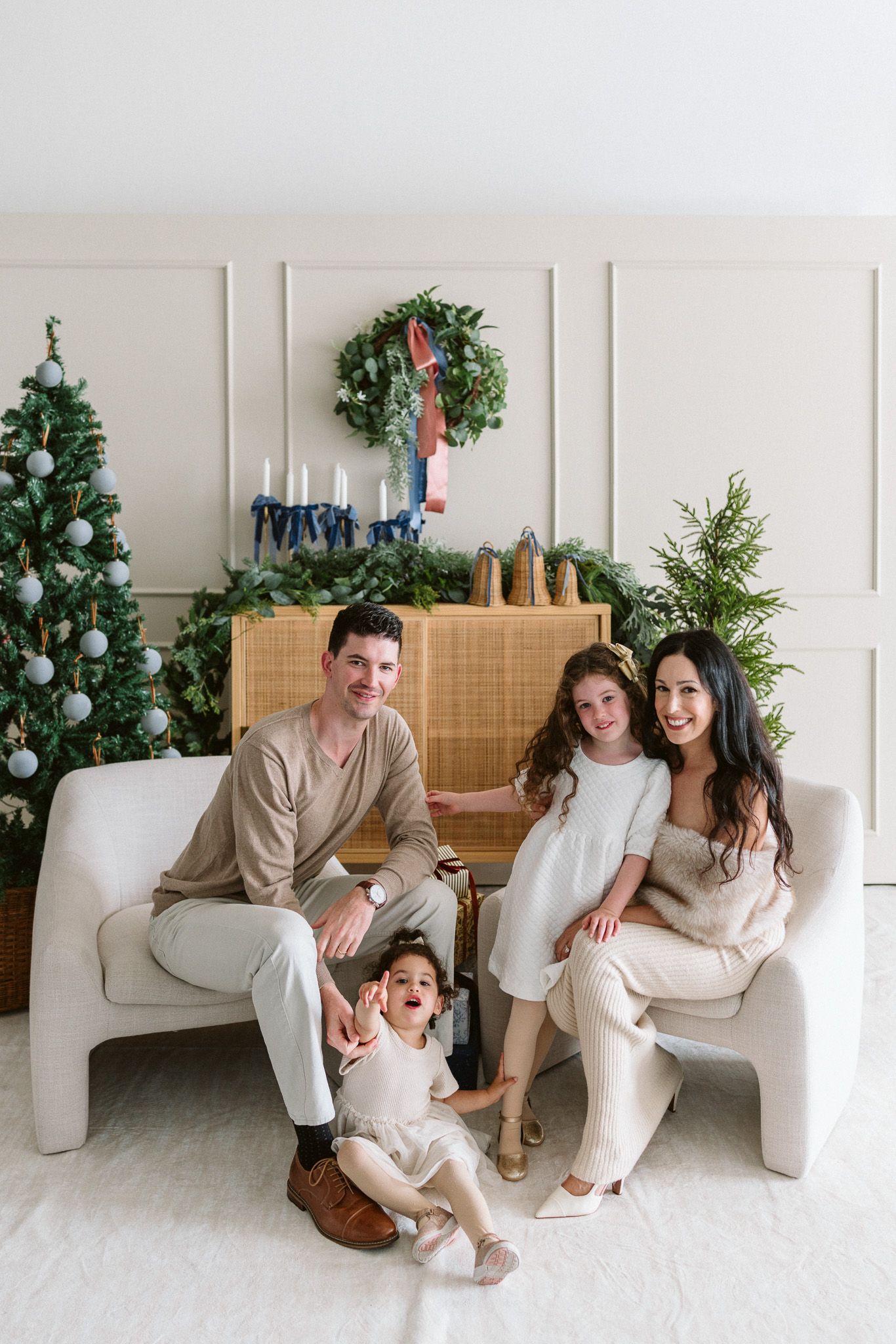 Family of four in cream outfits posing on white chairs beside a decorated Christmas tree, wreath, and candles in a bright holiday studio.