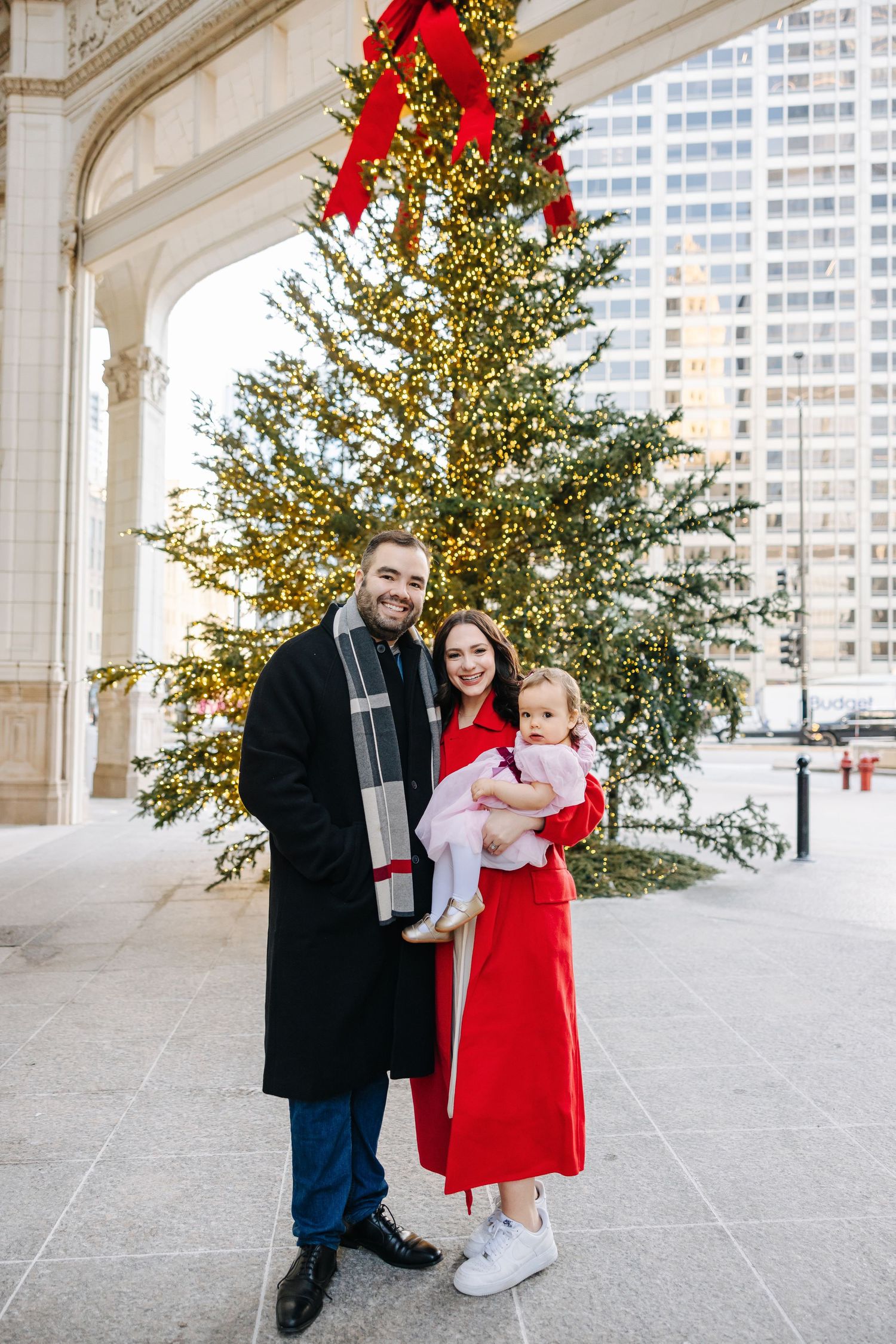Smiling family of three in winter coats poses with a toddler in front of a lit Christmas tree with a red bow near a city building.