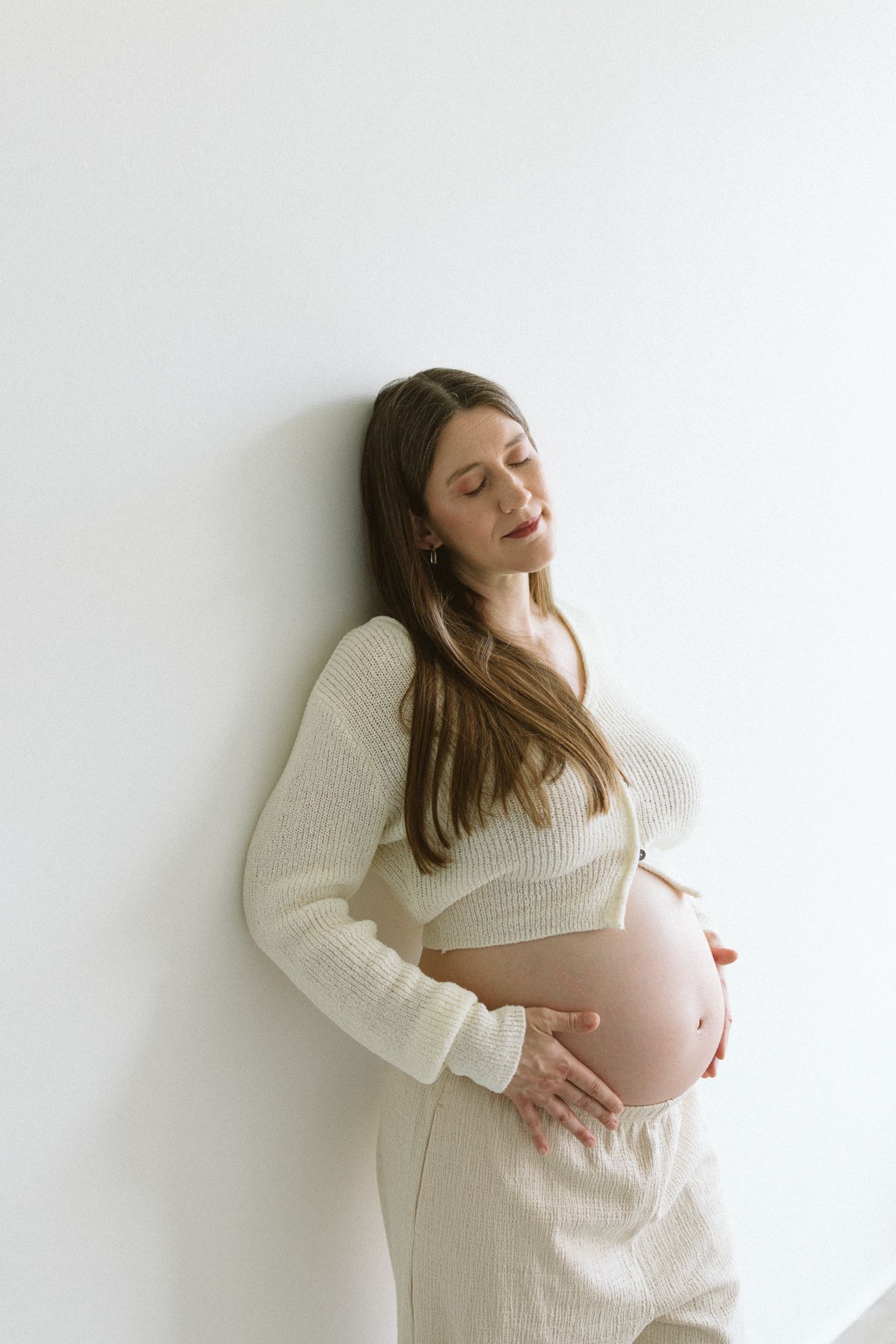 Expectant woman in cream knit sweater dress rests peacefully against white wall with hands cradling baby bump.