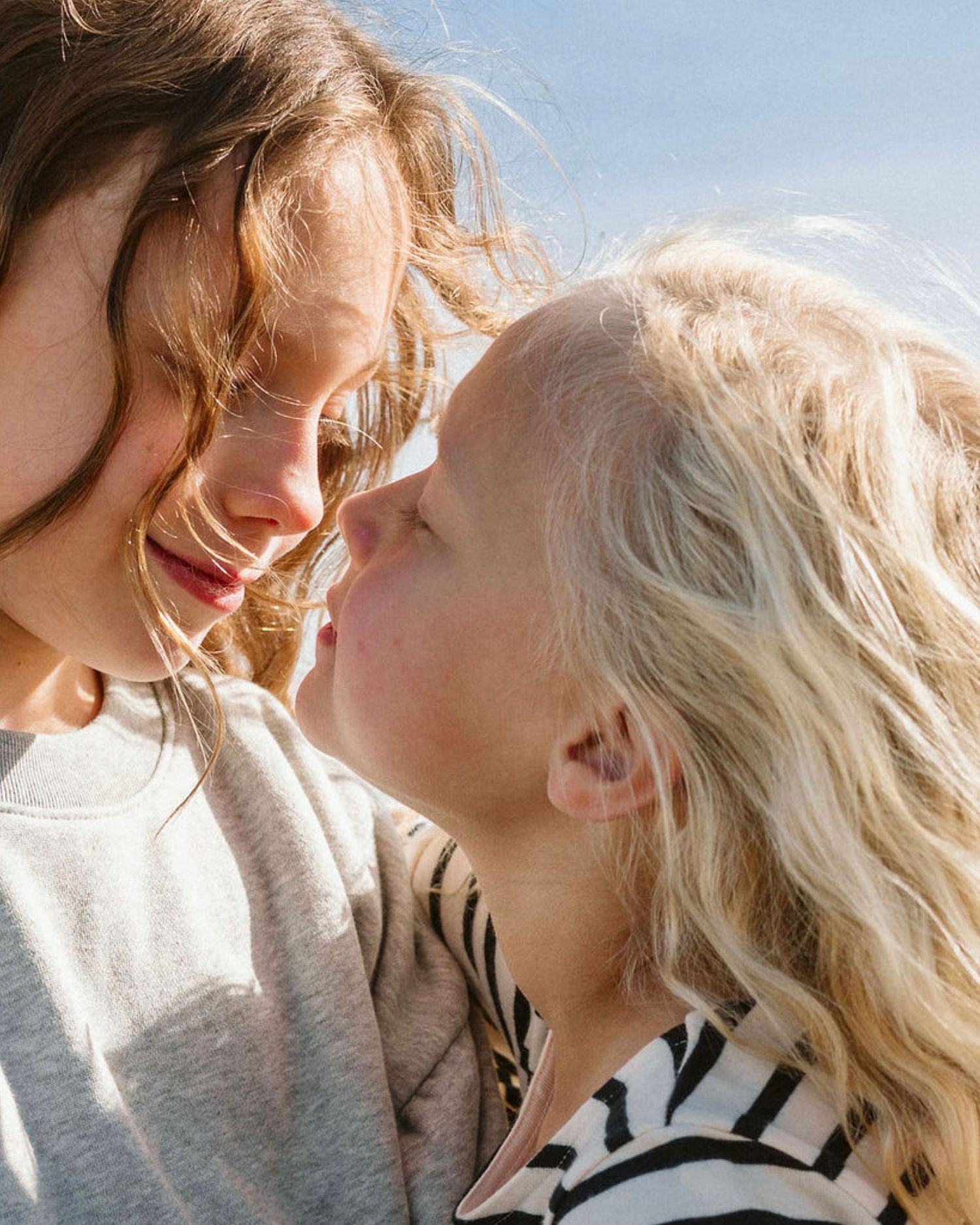 Two children with windswept hair share a tender nose-to-nose moment outdoors under a bright blue sky.