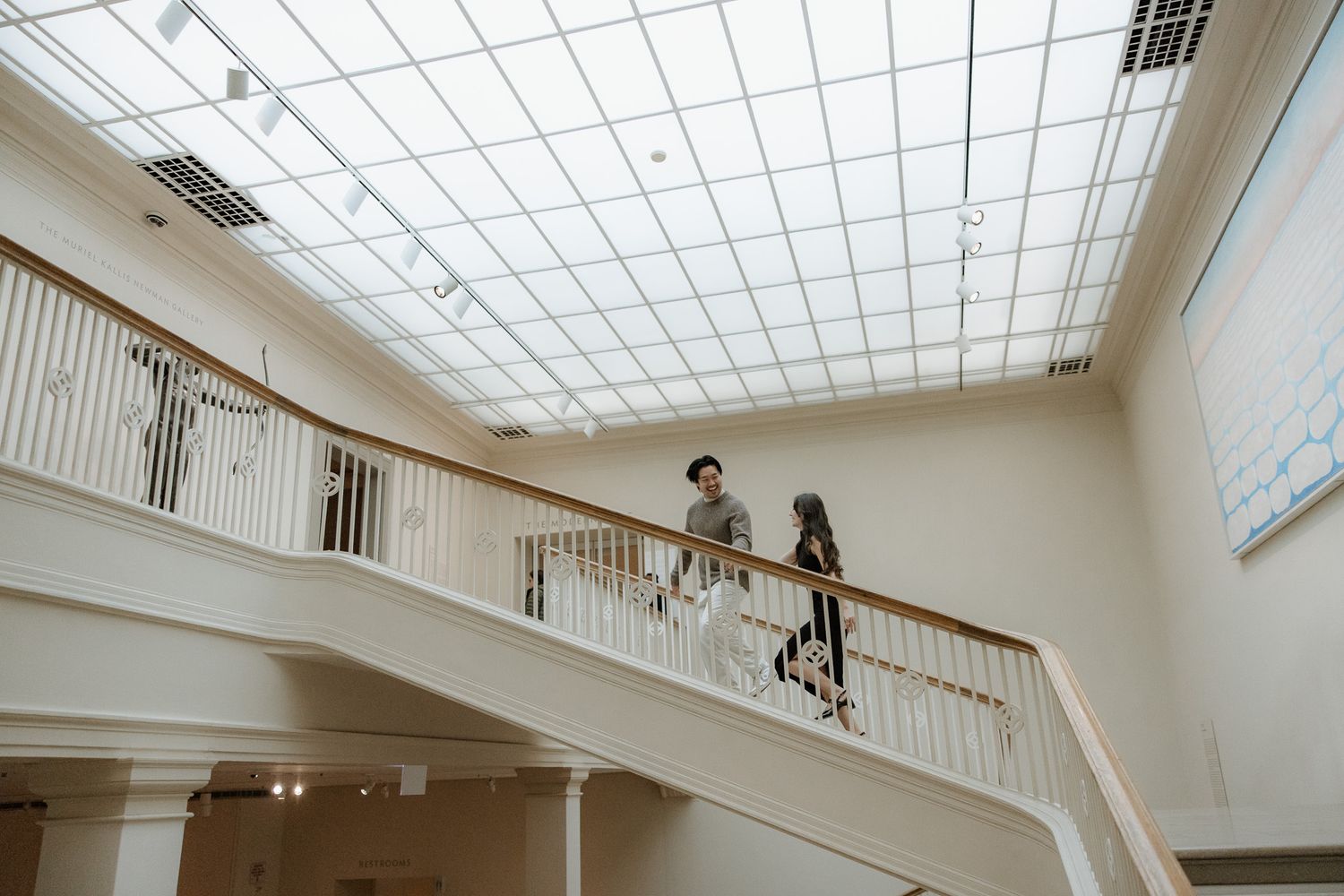 Two people descending a grand white staircase with skylight in an elegant interior.