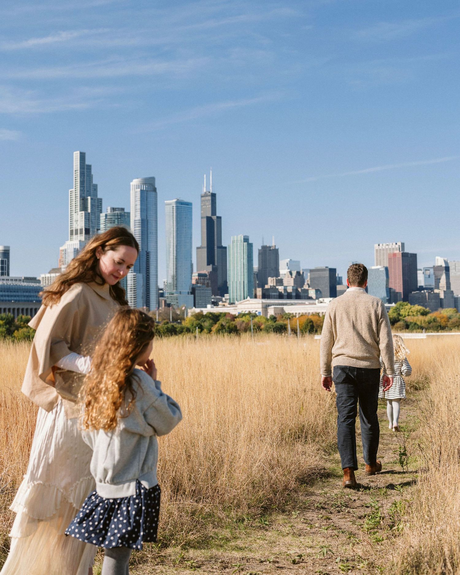 Family walking through golden prairie field with Chicago skyline in background on sunny day.