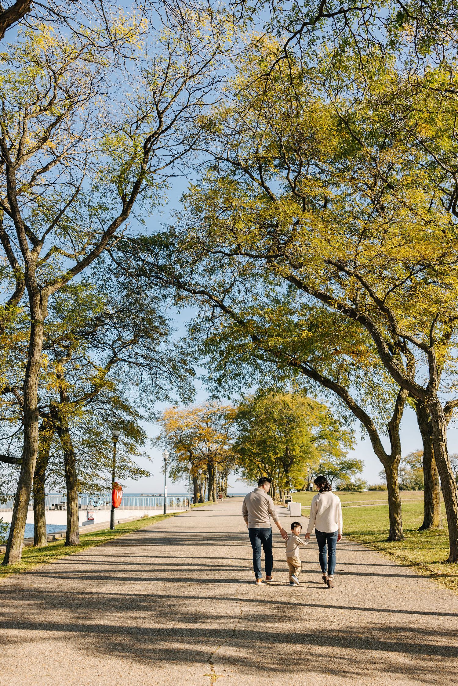 Family of three walking hand-in-hand along a tree-lined park path on a sunny autumn day near a waterfront.