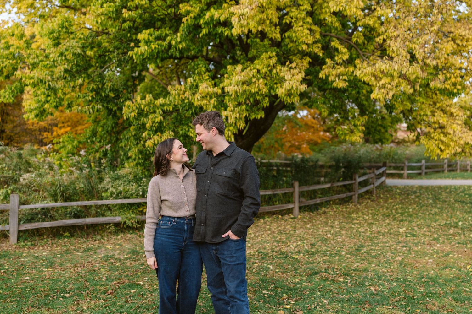 Smiling couple standing together on a grassy farm with a wooden fence and autumn trees.
