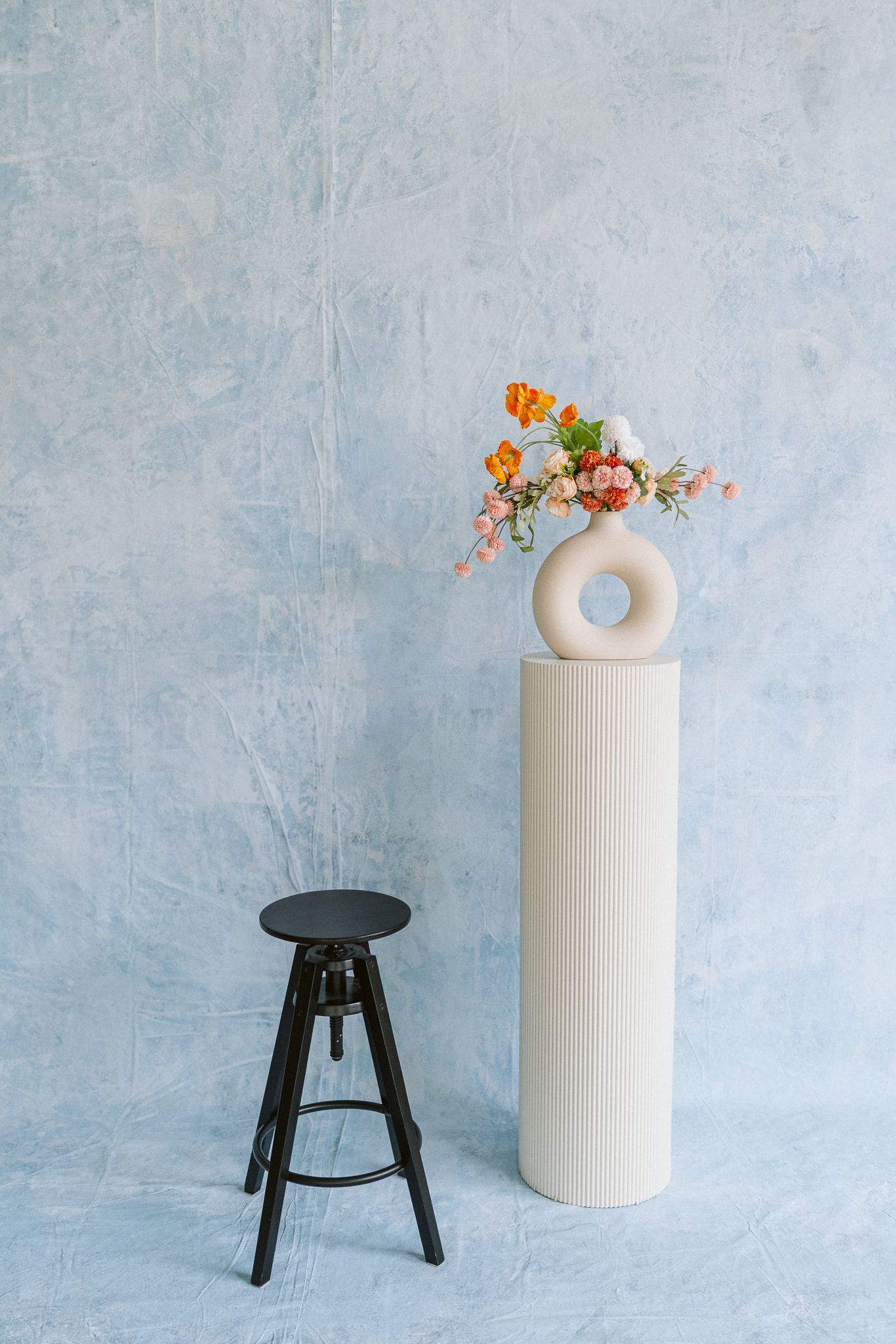 Minimalist still life series showing black stool and white ceramic vase with orange flowers against light blue wall.