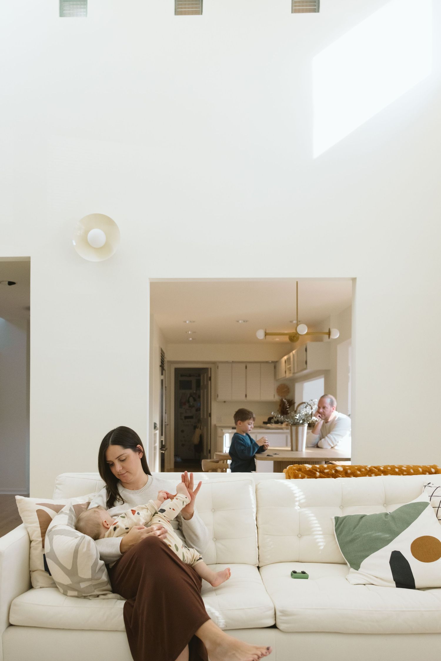 A bright, modern living room with people relaxing on a white couch while others are in the kitchen.