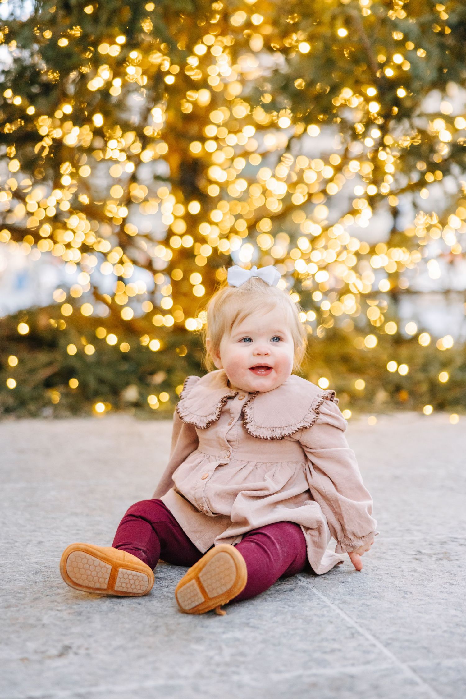 Smiling toddler in a blush ruffled jacket, burgundy pants and white hair bow sits outdoors before a tree glowing with golden string lights.