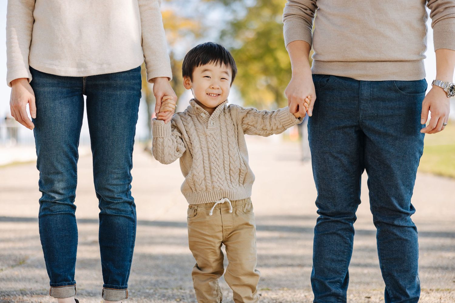 A smiling young Asian boy holds hands with his parents while walking outdoors in a park during autumn.