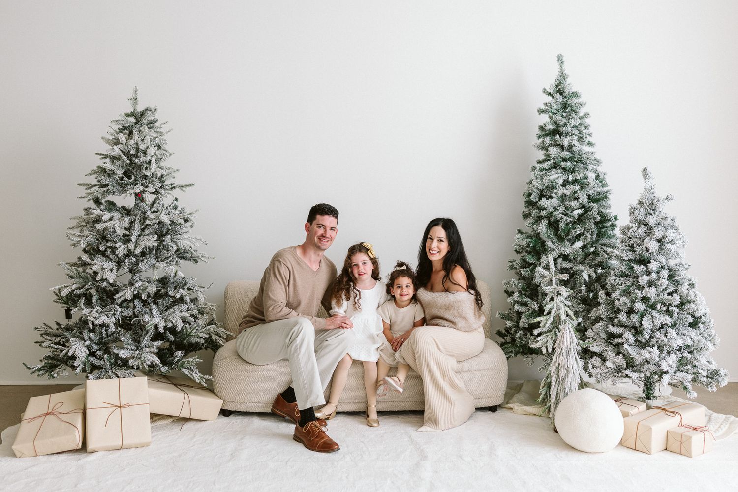 Family of four in neutral outfits sit on a cream sofa surrounded by flocked Christmas trees and wrapped gifts in a bright holiday studio.