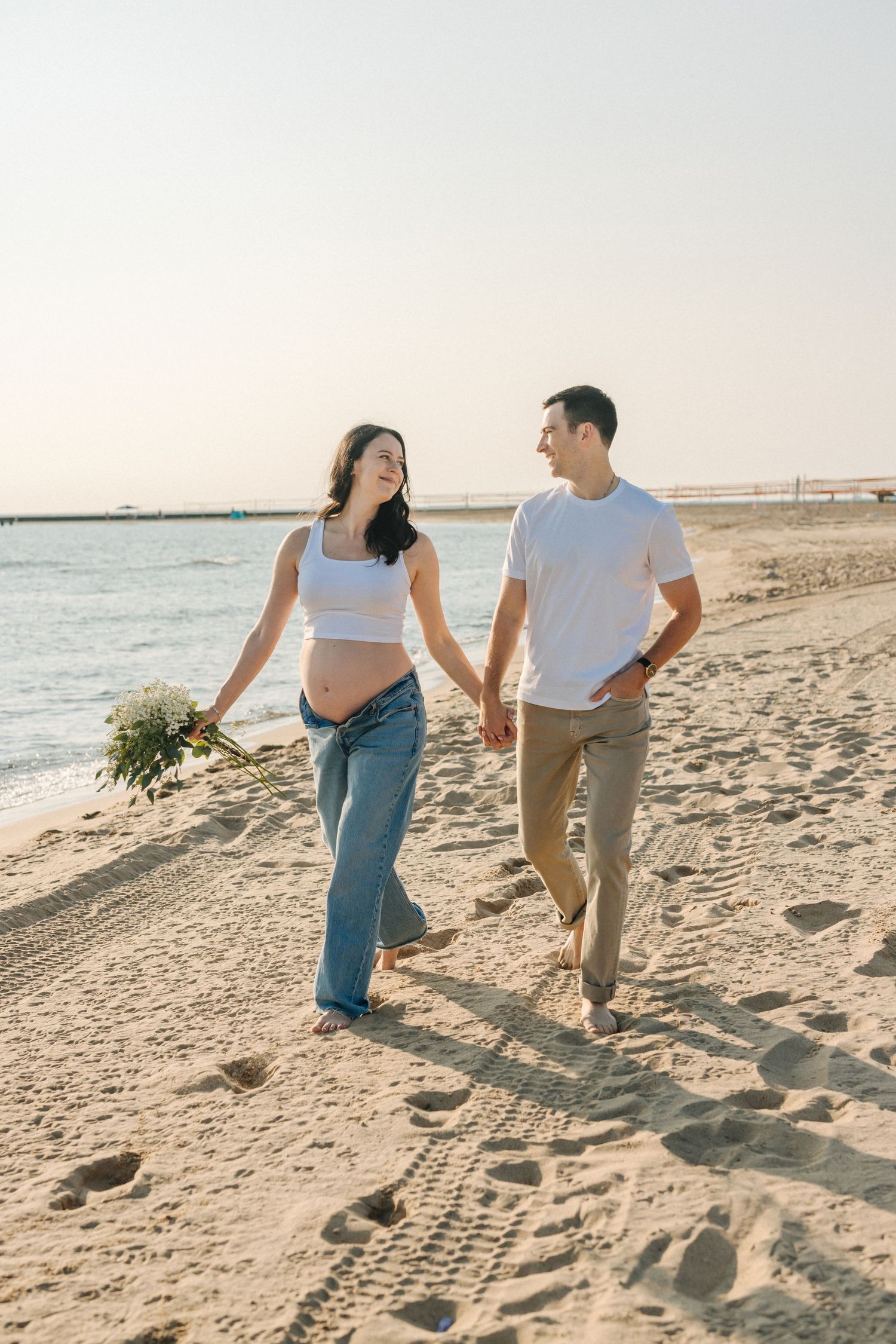 Couple walking hand in hand along sandy beach shoreline during sunset photoshoot.