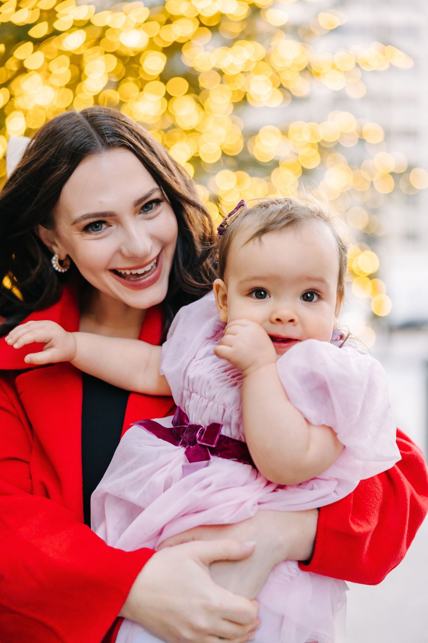Smiling woman in a red coat holds a baby girl in a pink tulle dress against a glowing golden bokeh of holiday lights.