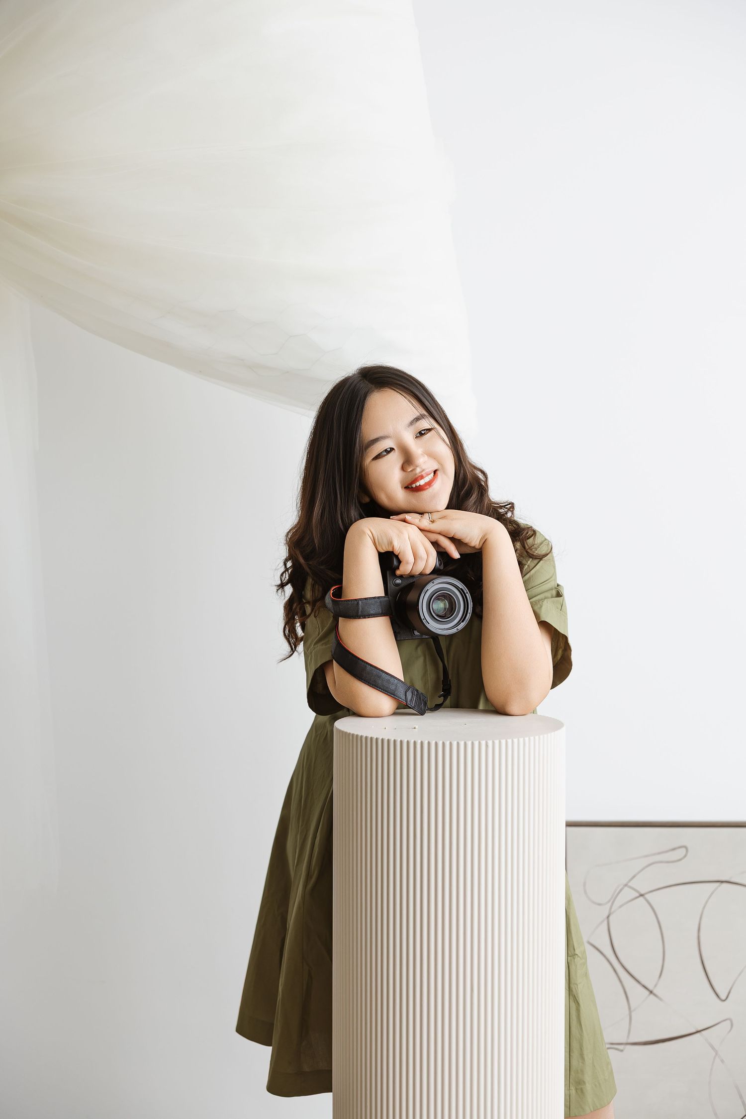 A photographer in an olive green dress poses with a vintage camera while leaning on a white column.
