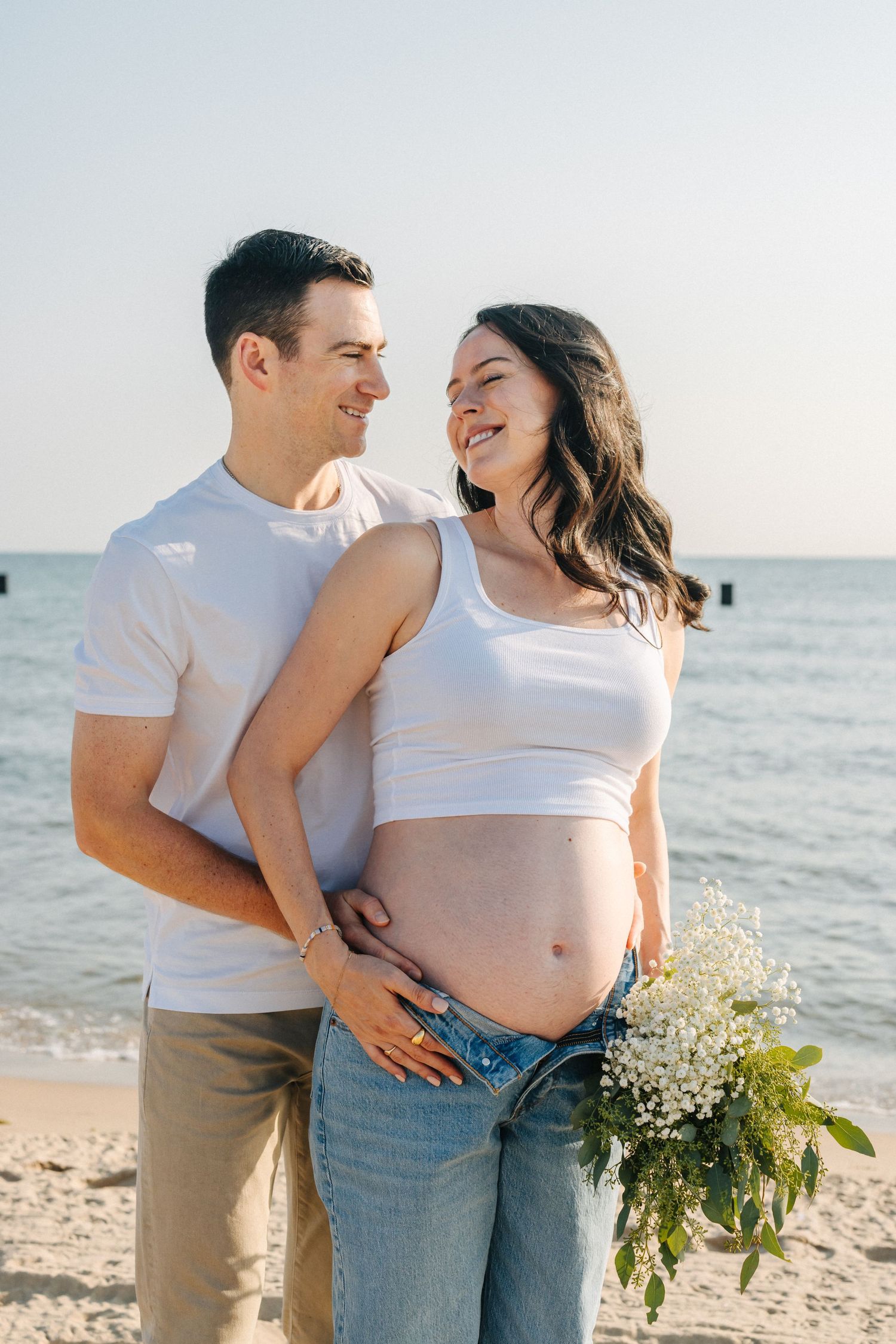 Expecting couple wearing matching white tops and jeans embrace on sandy beach with bouquet at sunset.