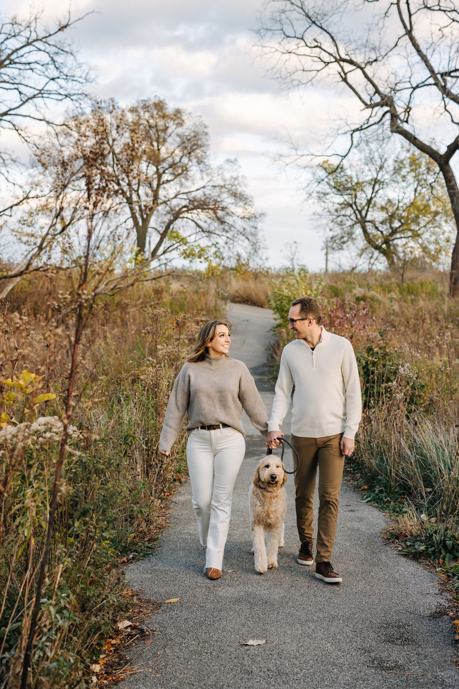 A couple walking their golden dog on an autumn path surrounded by bare trees and dry grass.