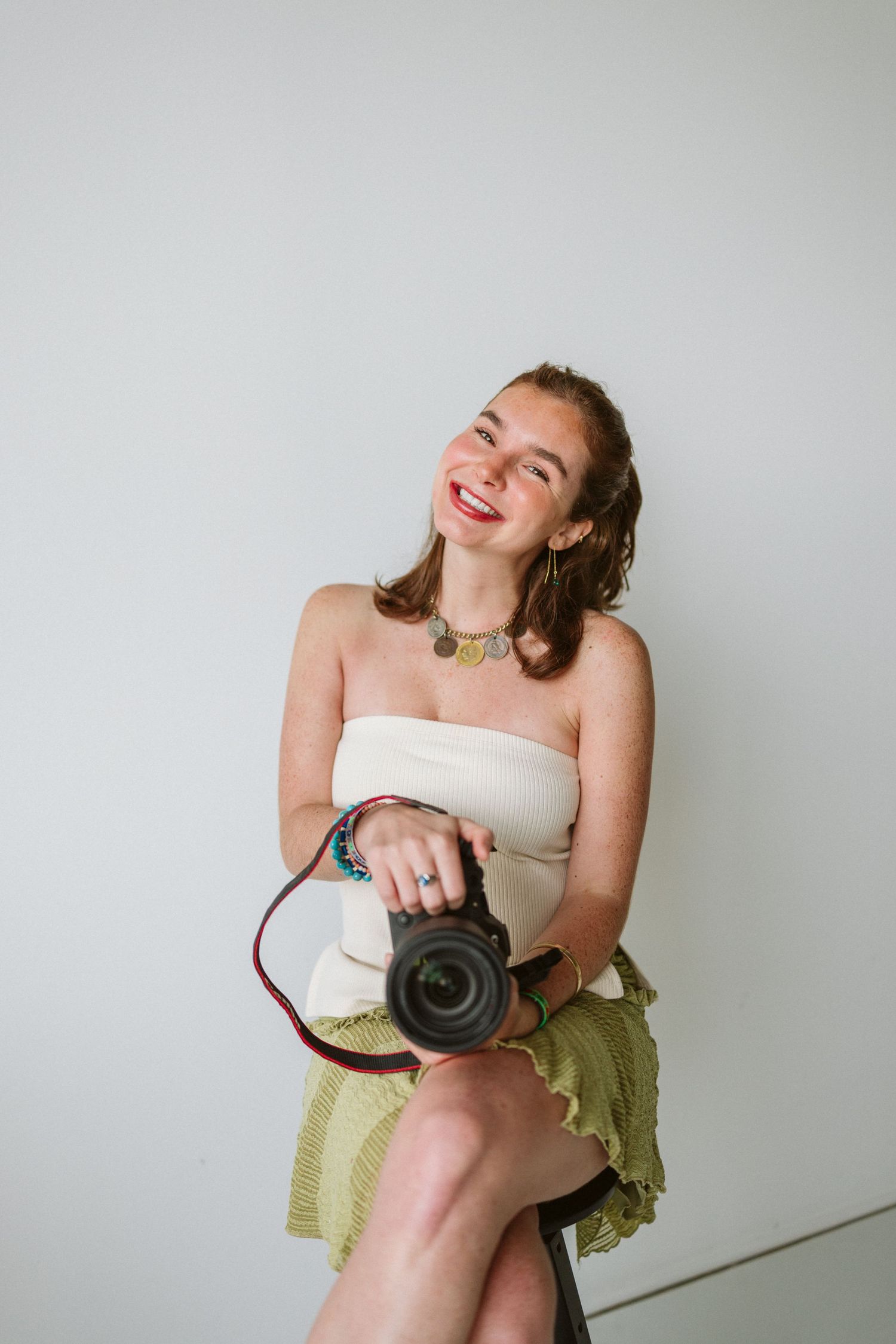 Person sitting casually in white top and yellow skirt while holding a camera and laughing against white wall.