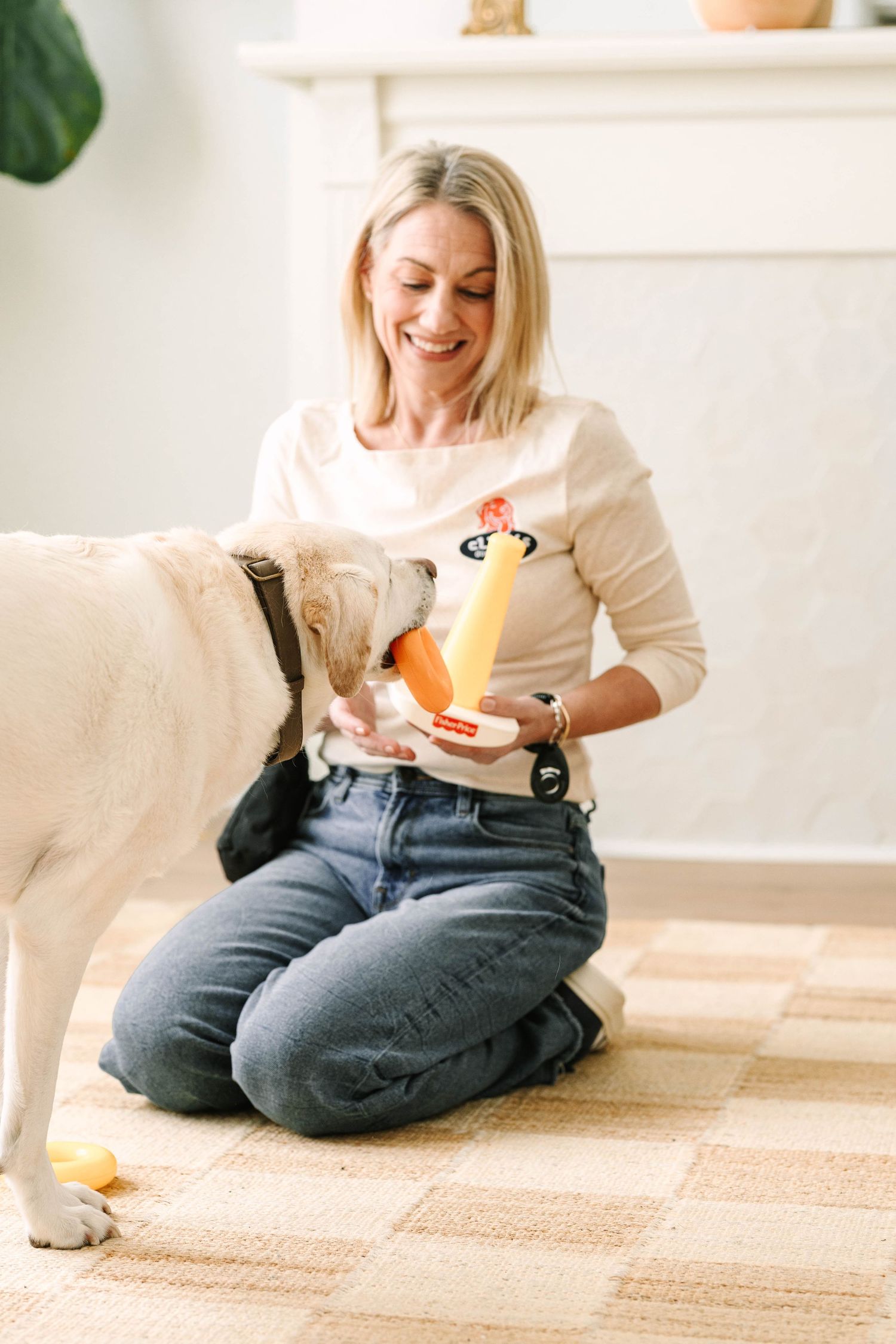 Person in white sweater and jeans plays with a golden retriever dog on a carpeted floor while holding a toy.