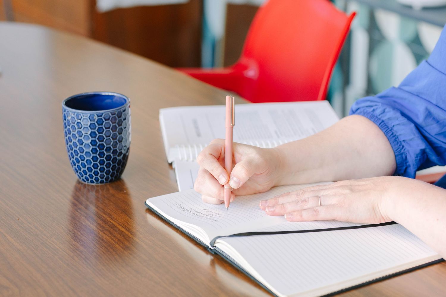 Person writing in open notebook with pencil beside blue ceramic mug on wooden table.