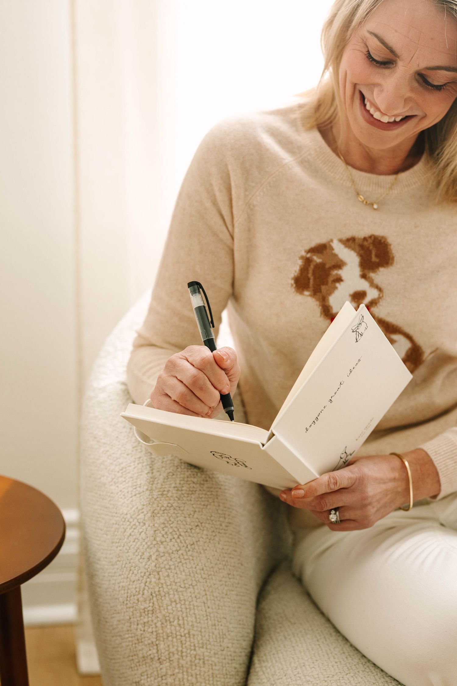 Person in cream sweater sitting on white couch writing in book with black pen while smiling.