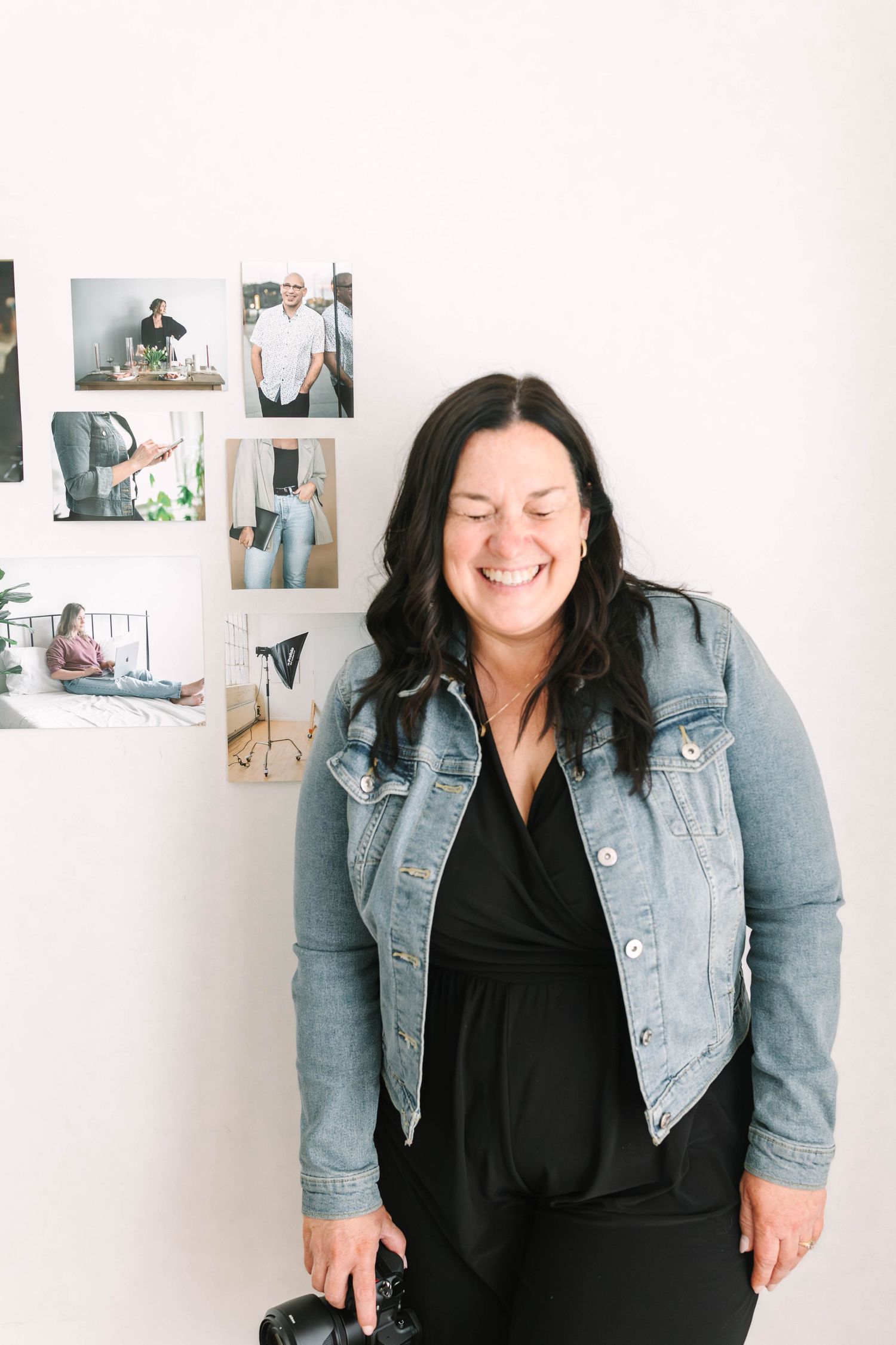 A person wearing a denim jacket and black dress smiles joyfully against a white wall with photos.