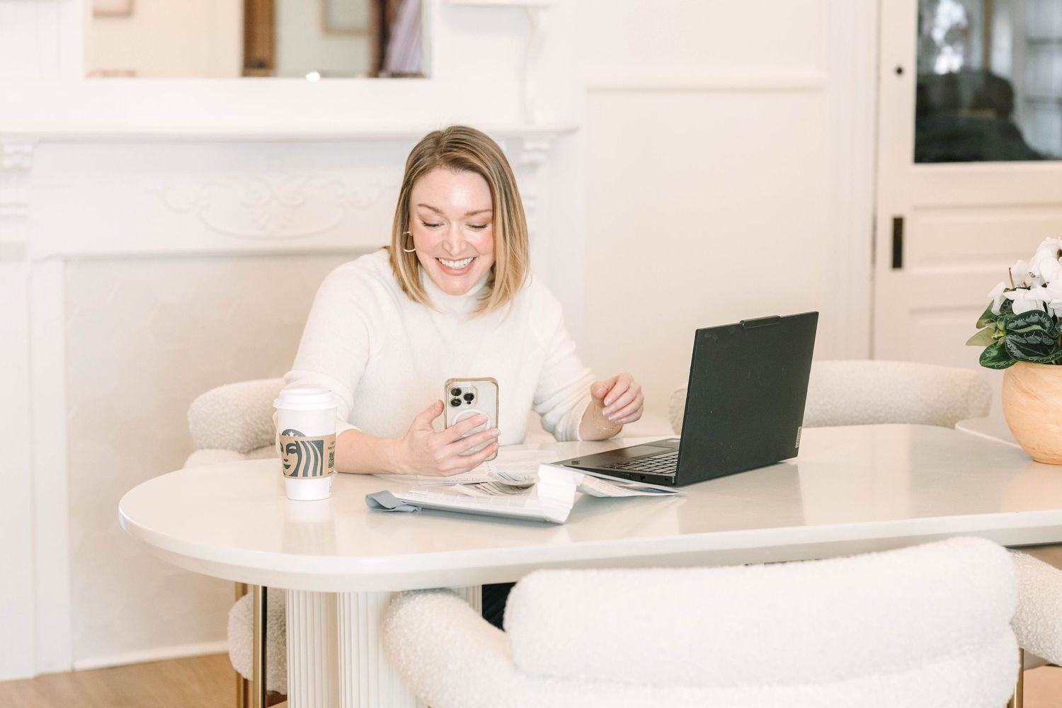 Professional working at white desk with laptop and smartphone in bright modern home office setting.