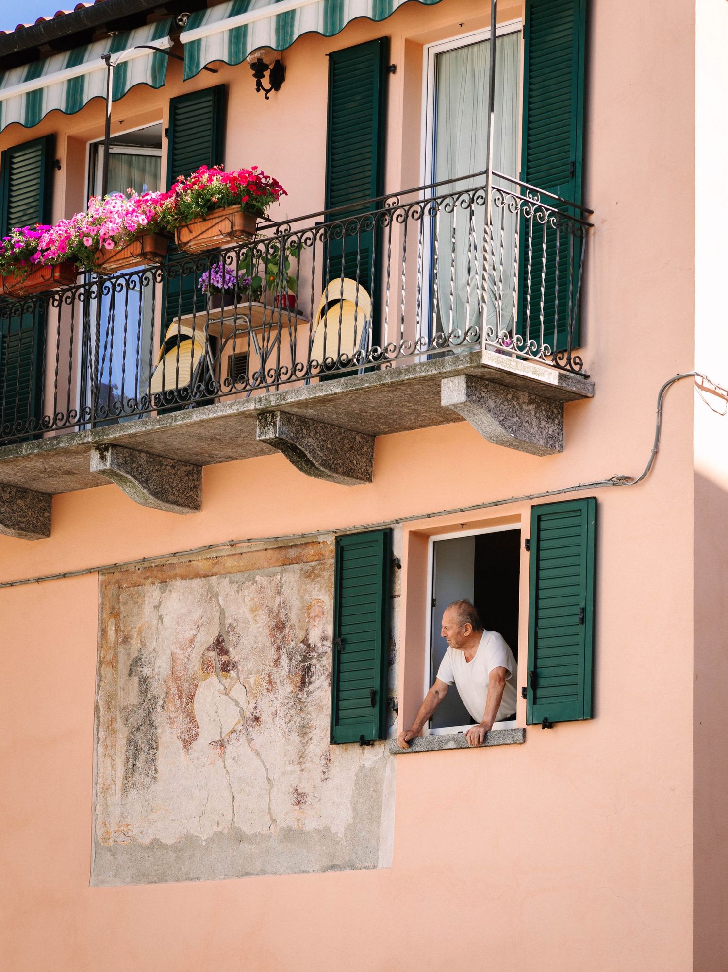 Man leaning out of open window with green shutters on a peach-colored Italian building with balcony and flowers.