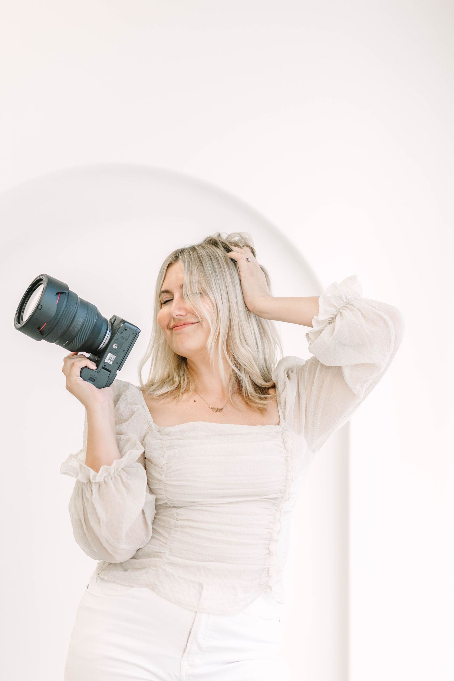 A photographer in a white dress poses playfully with a camera in a bright studio setting.