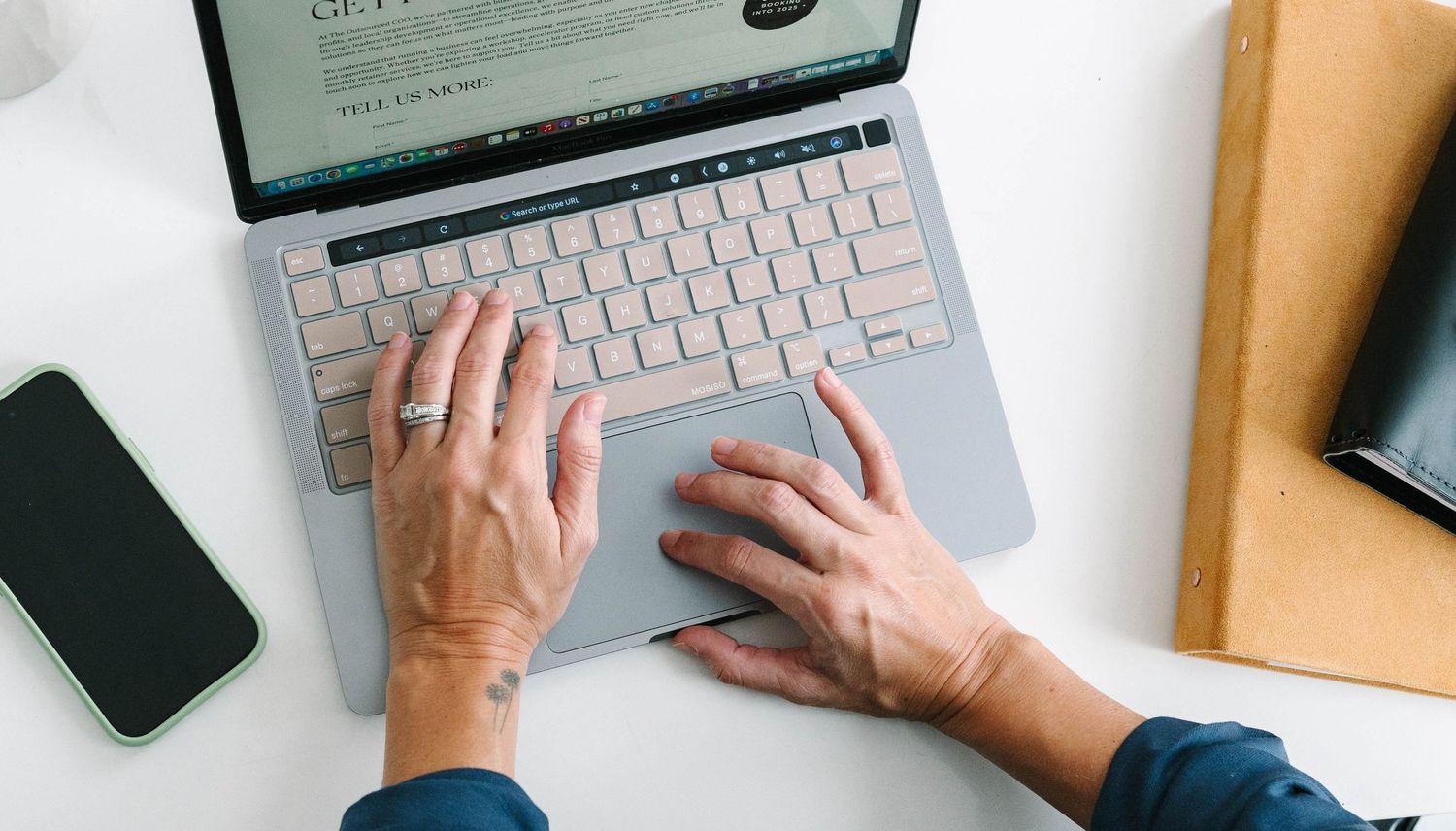 Overhead view of hands typing on laptop keyboard with phone and notebook on desk.