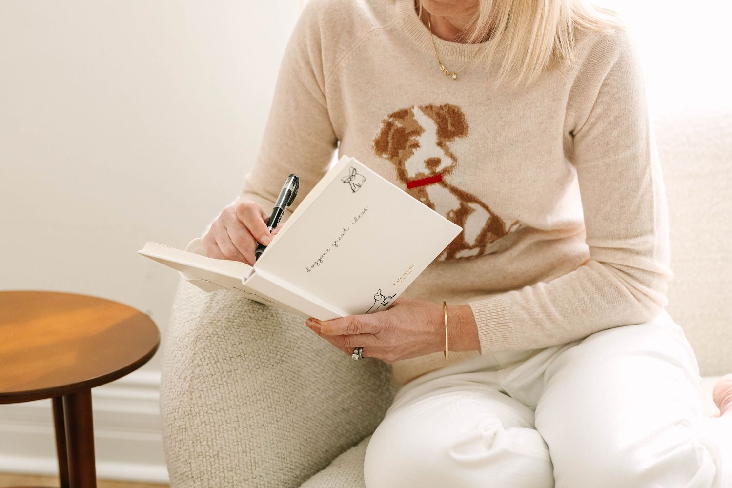 Person in white sweater sitting at wooden table writing in notebook while holding coffee mug.