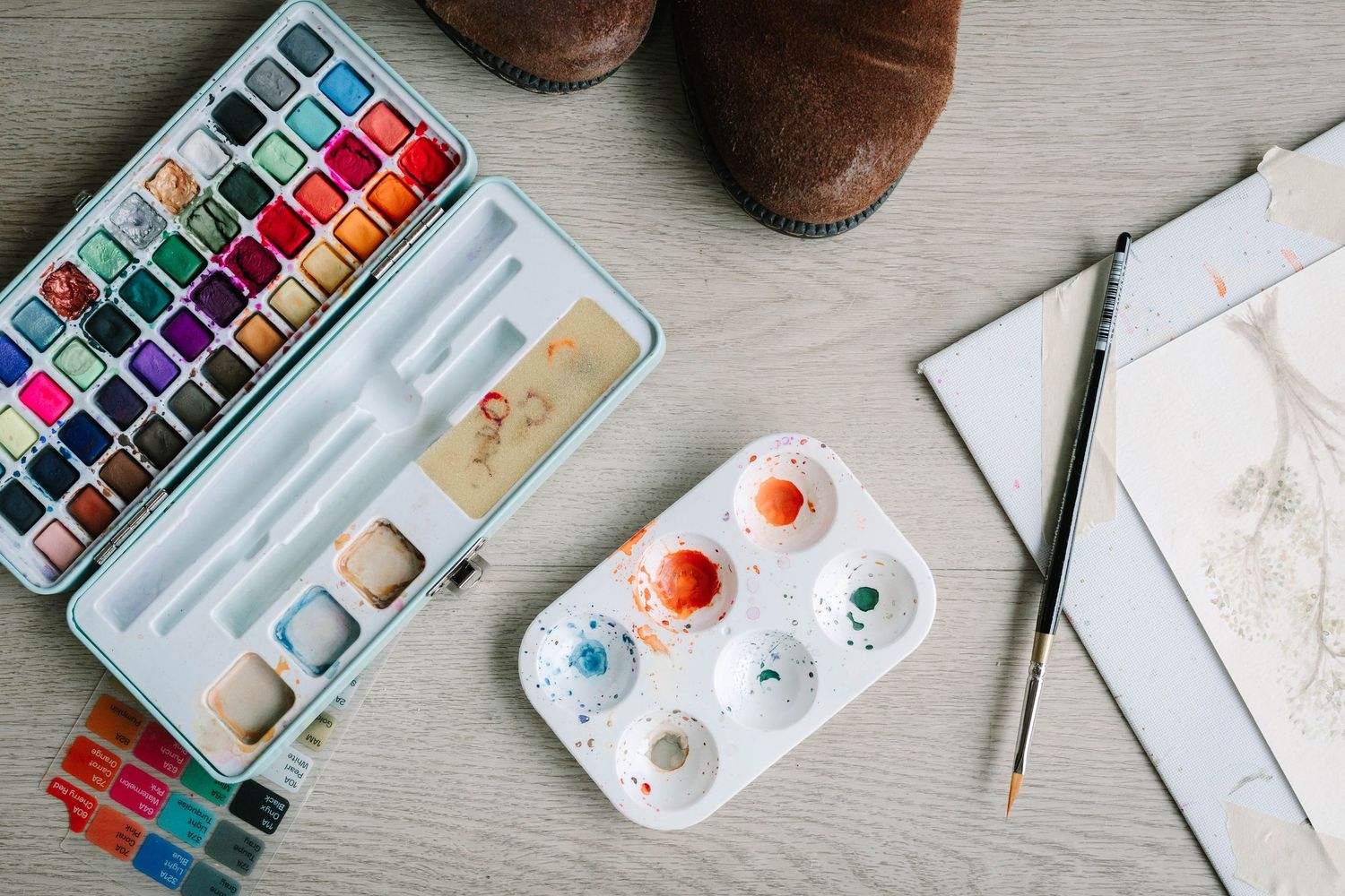 Flat lay of watercolor art supplies including paint palettes and brushes on light wood surface.