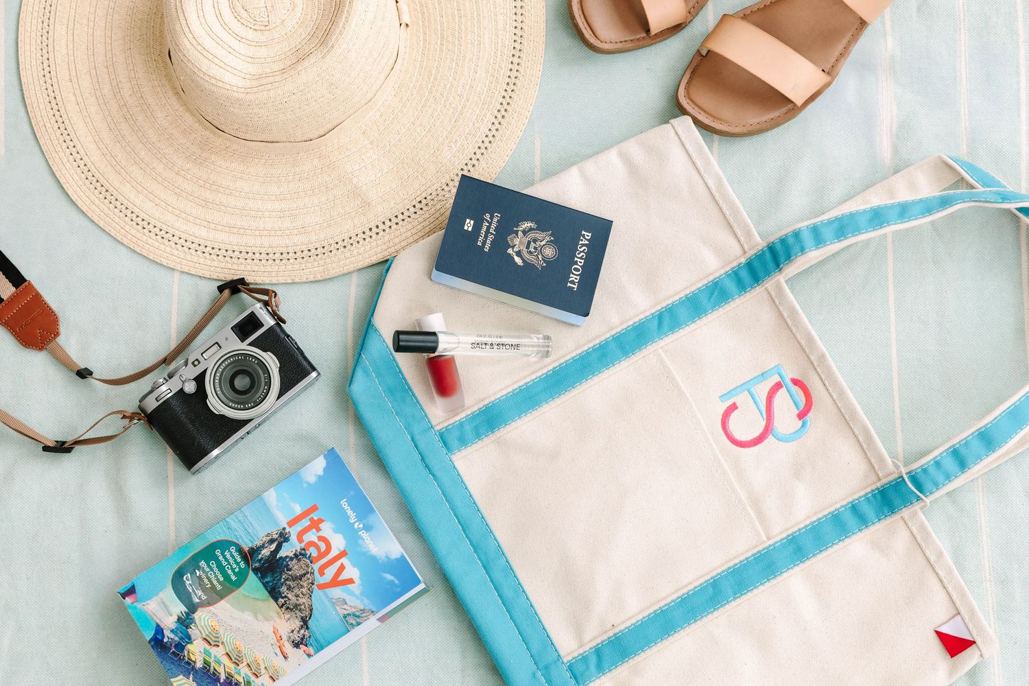 Flat lay of travel essentials including a straw hat, passport, camera, sandals, tote bag, and guidebook on a light blue surface.