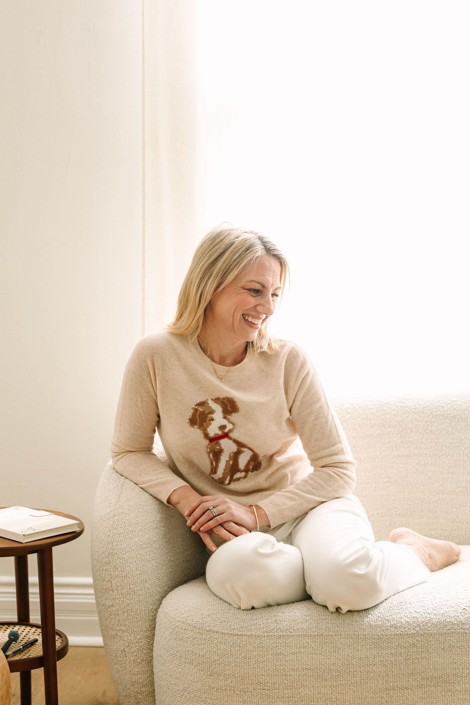 Woman in beige sweater relaxing and playing with small brown and white puppy on white couch.
