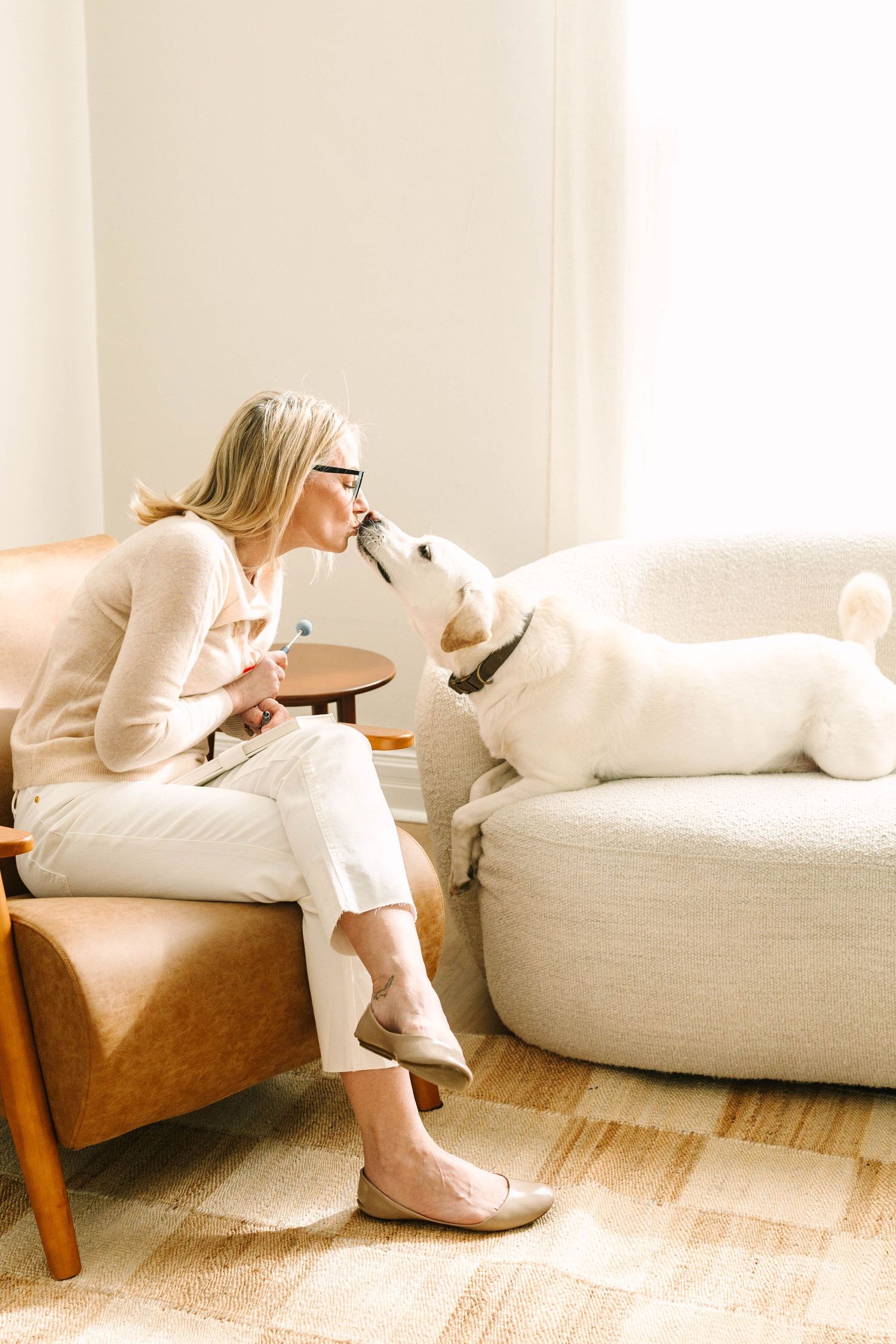 A white Labrador dog and someone in cream-colored clothing share an affectionate moment on a cozy living room couch.
