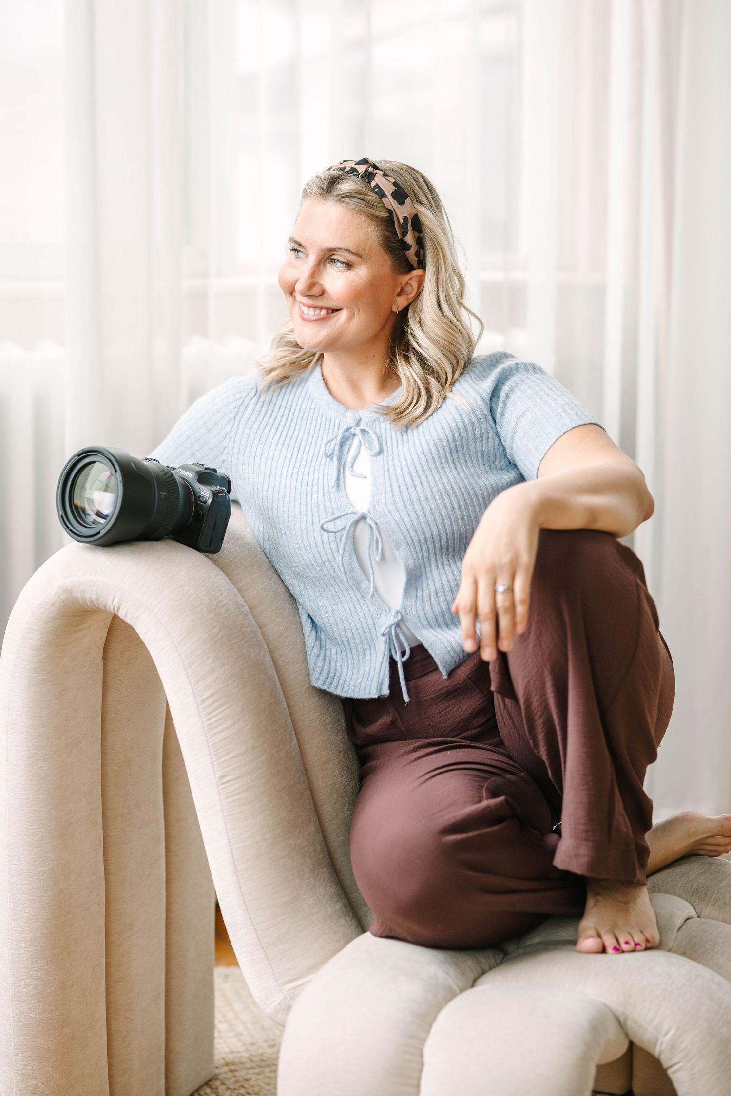 Pregnant woman in blue shirt relaxes on beige couch by window with natural light streaming in.