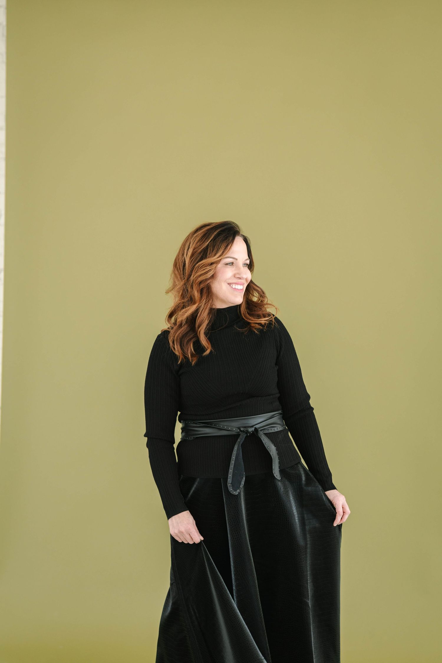Woman in black turtleneck and satin skirt smiling against olive green wall.