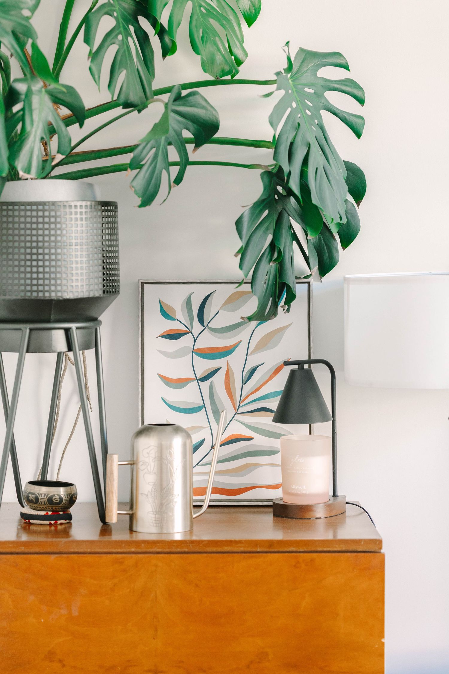 Wooden dresser styled with a candle lamp, silver vase, botanical art print, and large monstera plant in a modern boho interior.